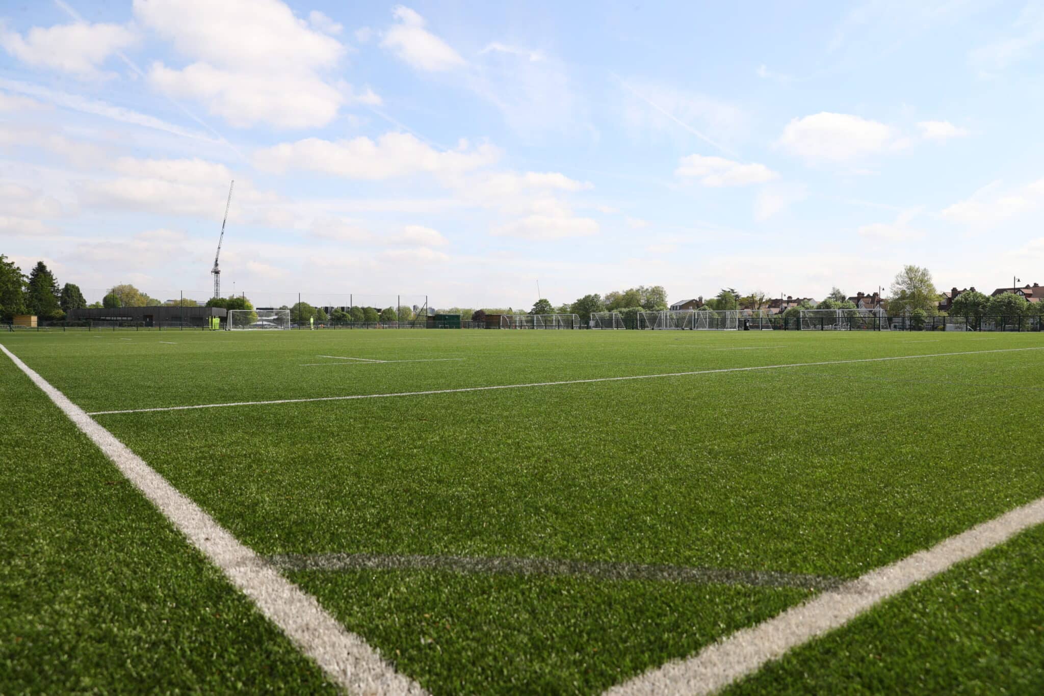 A wide angle view of an empty, green sports field with white lines, under a partly cloudy sky. Trees and buildings are visible in the distance.