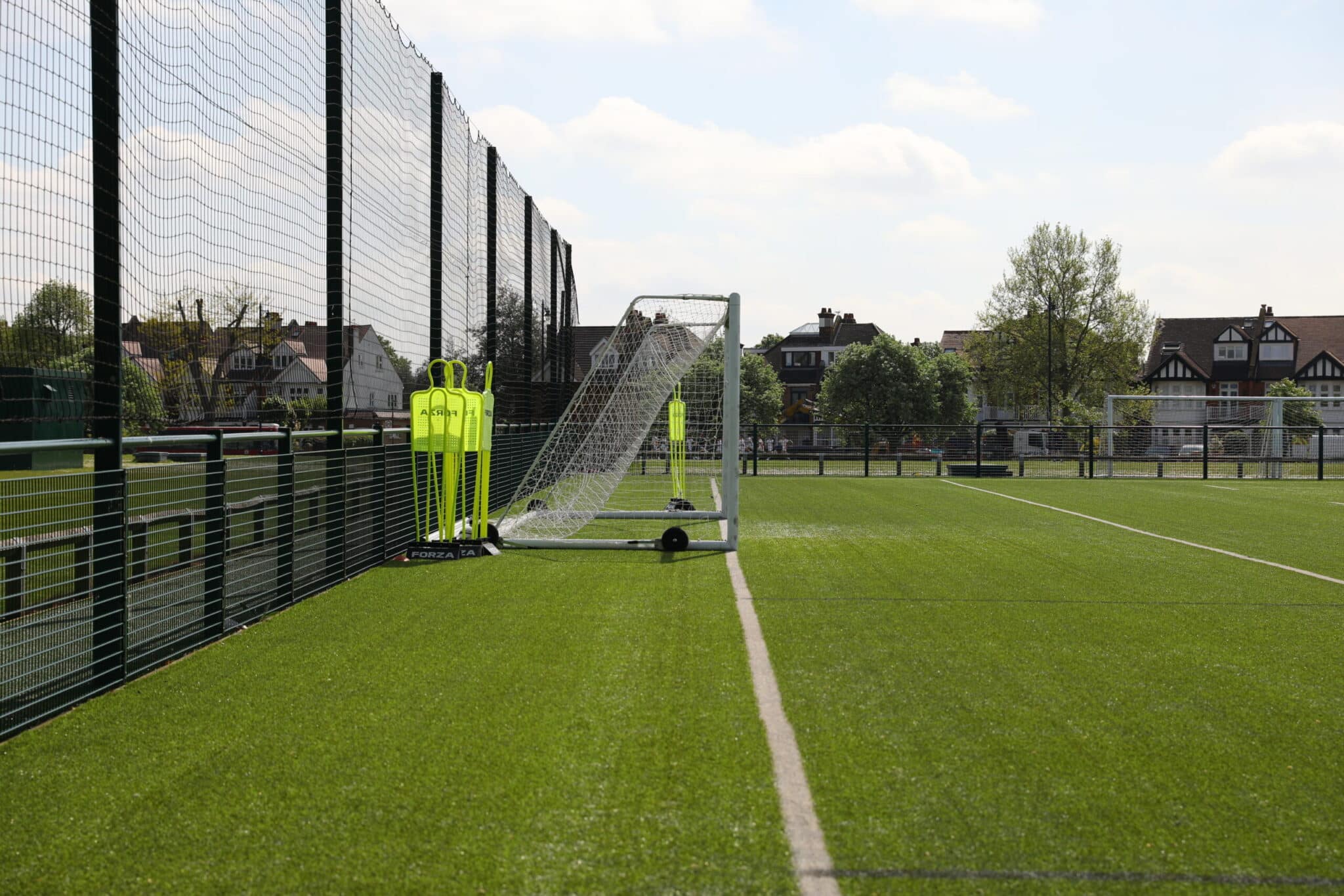 A soccer goal and practice mannequins are positioned on the edge of a green artificial turf field, next to a tall fence, with houses and trees visible in the background under a partly cloudy sky.