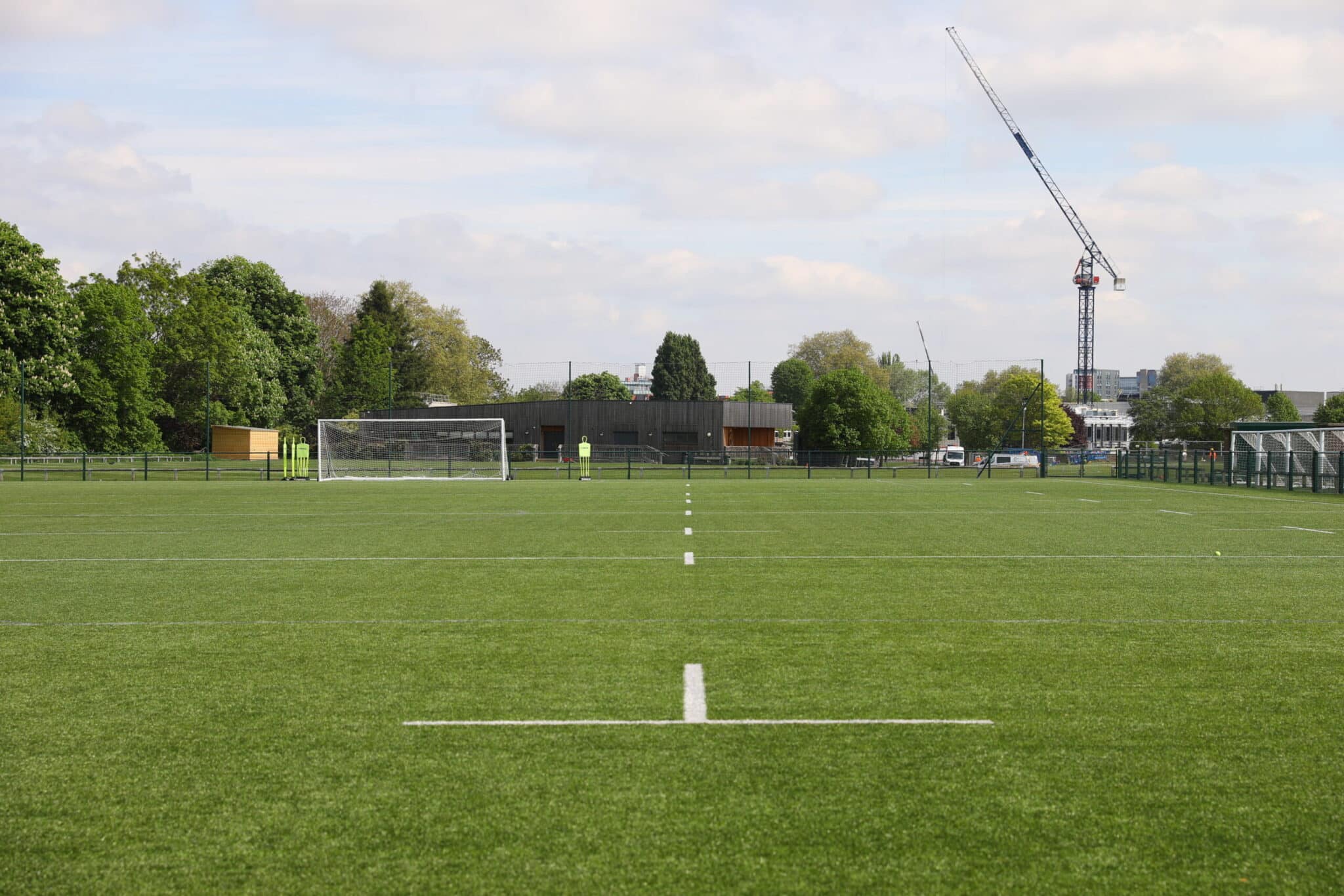 A green, empty sports field with white lines, a goalpost at the far end, trees and buildings in the background, and a tall construction crane visible under a partly cloudy sky.