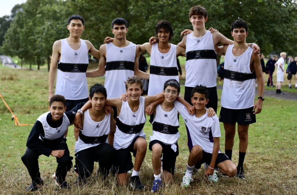 A group of ten boys wearing matching athletic uniforms pose together outdoors on grass, smiling after a Cross-Country race. Trees and other people can be seen in the background.