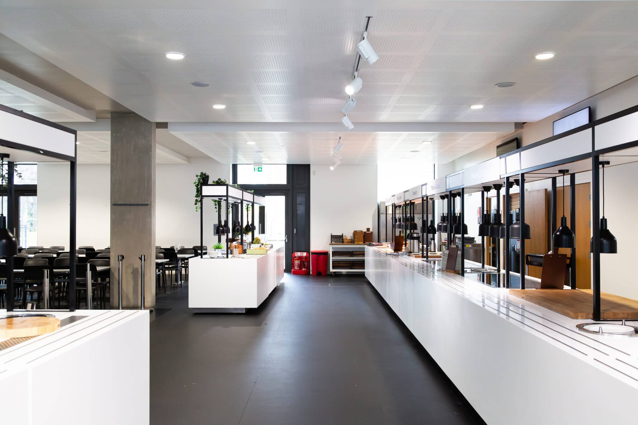 A modern, empty cafeteria with long white serving counters, black lamps, and a dining area with black chairs and tables in the background. The space is brightly lit with natural and ceiling lights.
