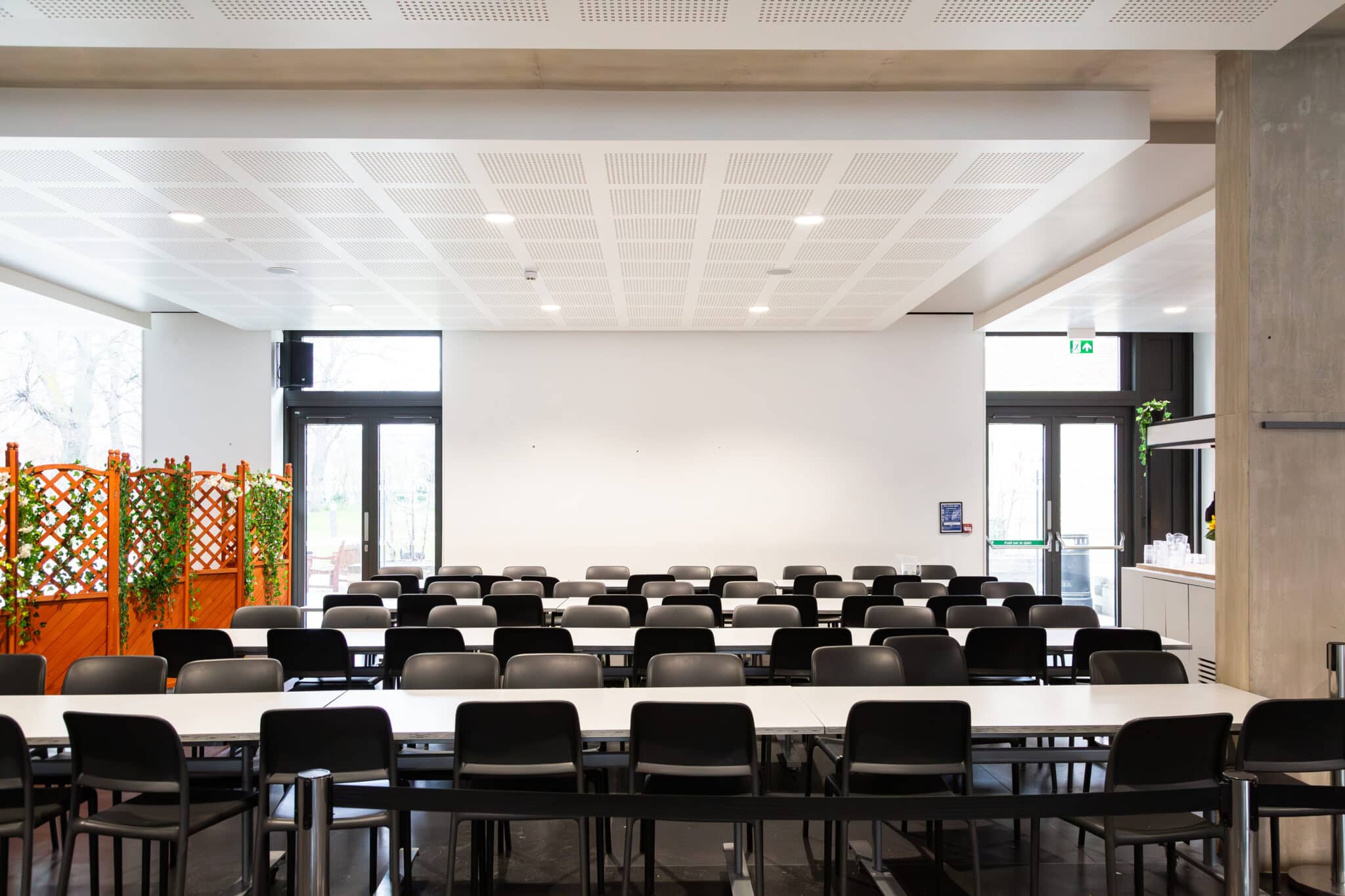 A modern, empty cafeteria with rows of black chairs and white tables, large windows letting in natural light, and some green plants in orange planters on the left side.