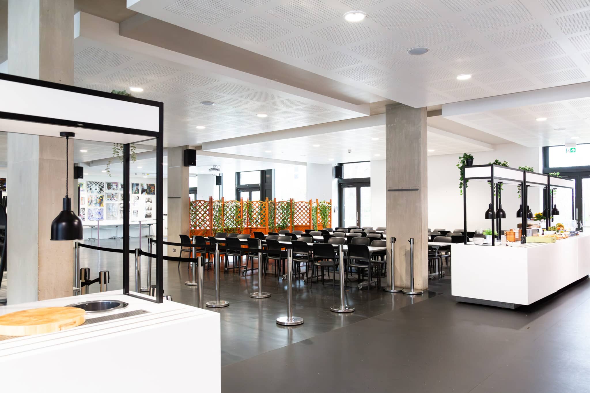 A modern, spacious cafeteria with rows of empty black chairs and tables, food serving counters, and bright lighting. There are dividers with a colorful pattern and large windows letting in natural light.