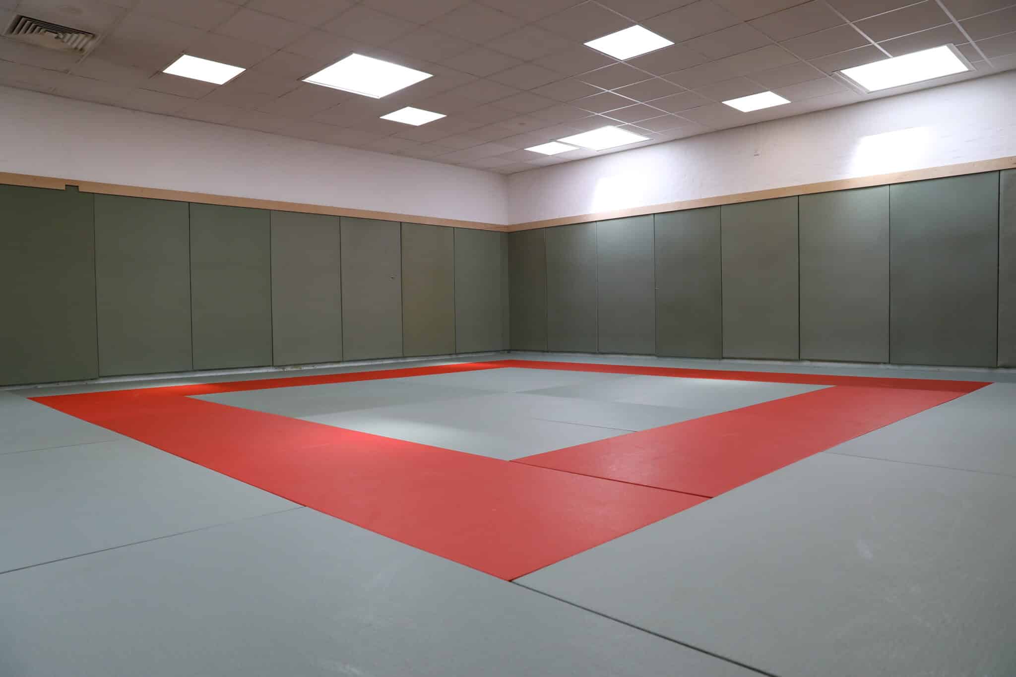 A spacious, empty martial arts dojo with padded gray and red mats covering the floor and soft green wall panels, illuminated by overhead fluorescent lights.