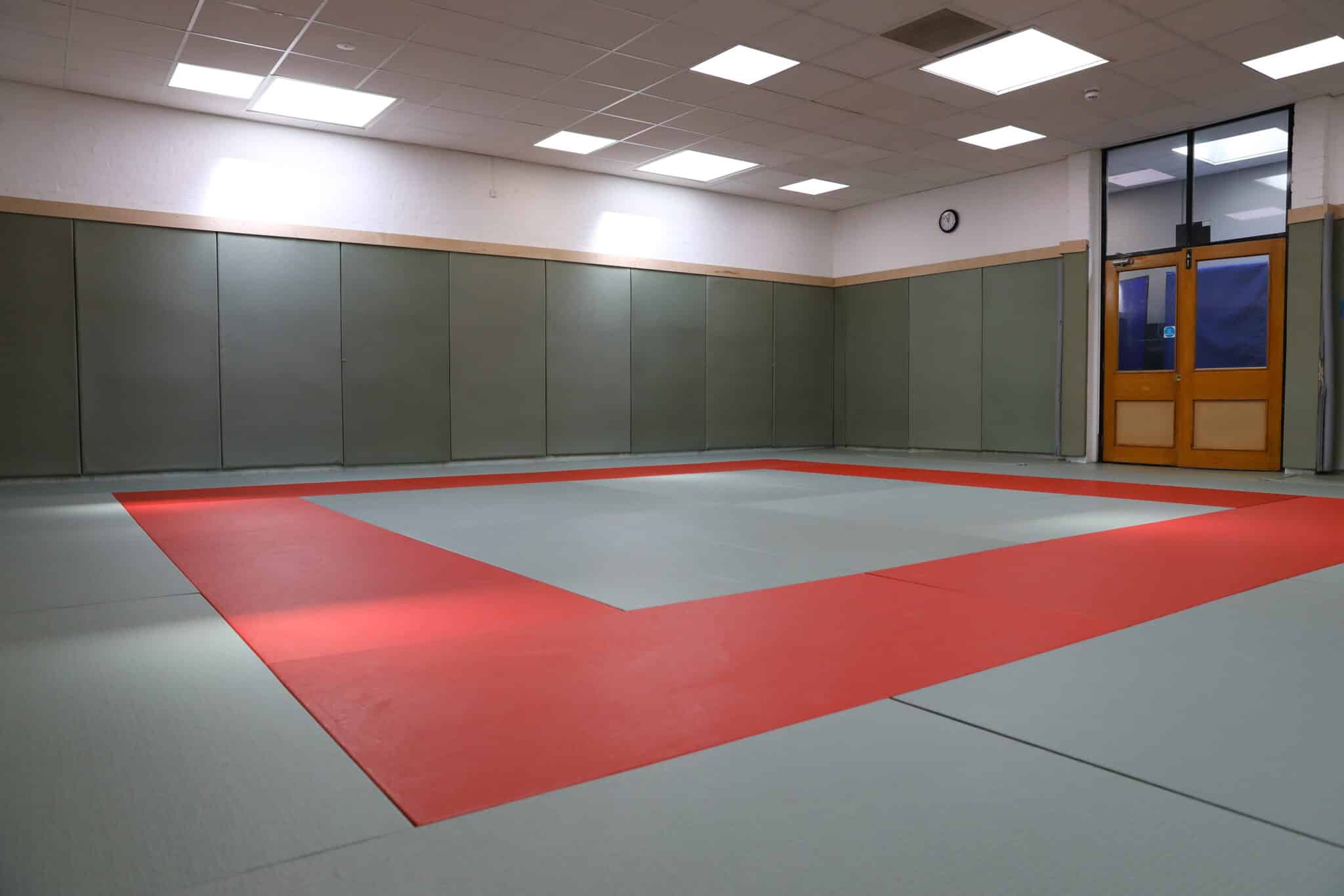An empty martial arts dojo with padded green walls, a red and gray mat on the floor, fluorescent ceiling lights, a clock on the wall, and a door with glass windows.
