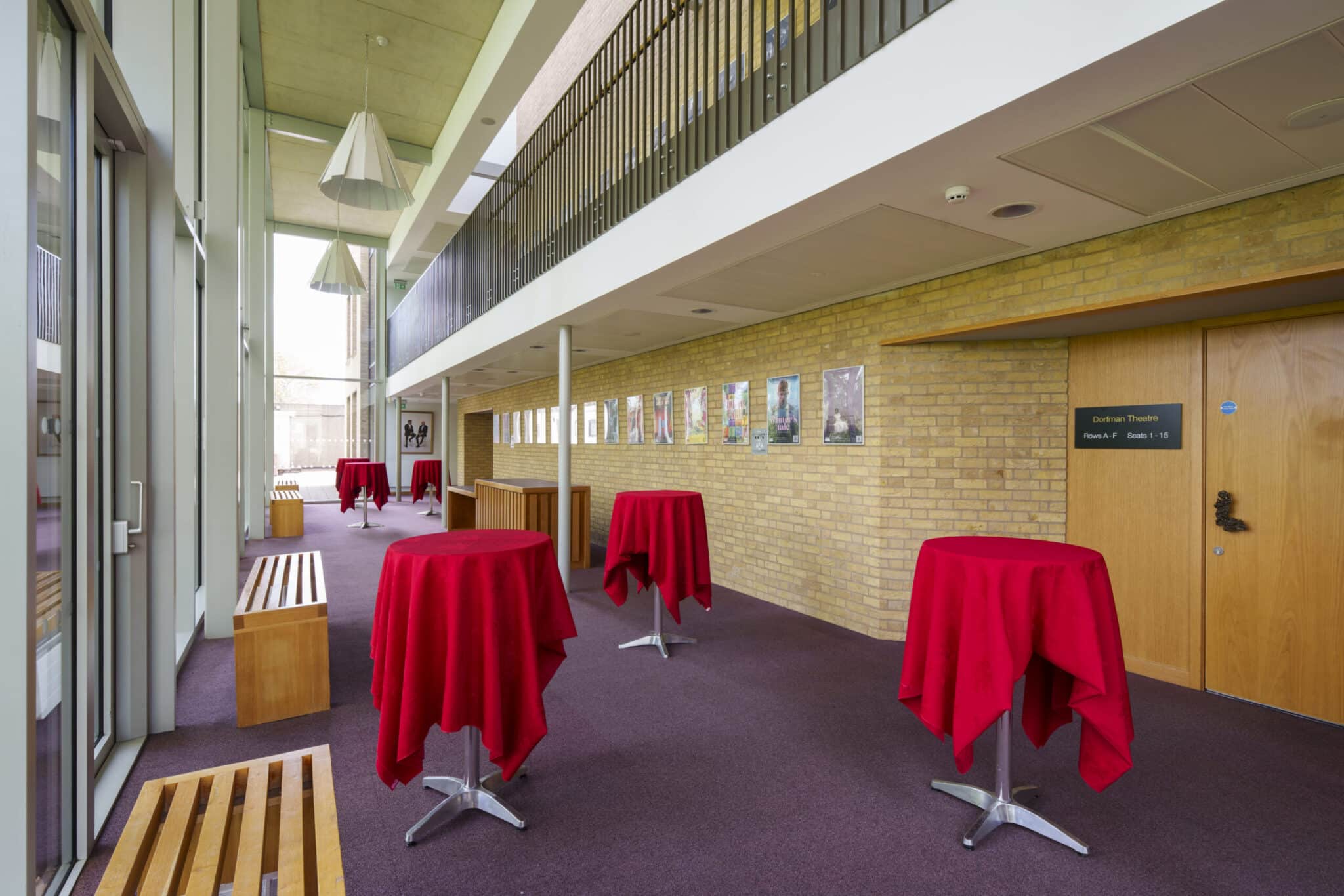 A modern hallway with tall windows, wooden benches, and high tables covered with red tablecloths. The walls are lined with framed photos, and there are doors and signs along the right side.