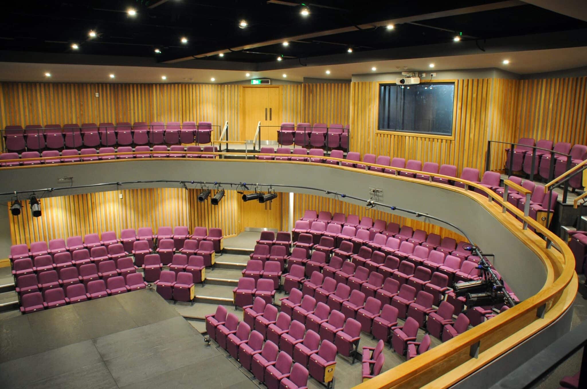 A modern theater auditorium with tiered rows of purple seats, wooden wall panels, a balcony section, and stage lighting equipment; the stage area is empty.