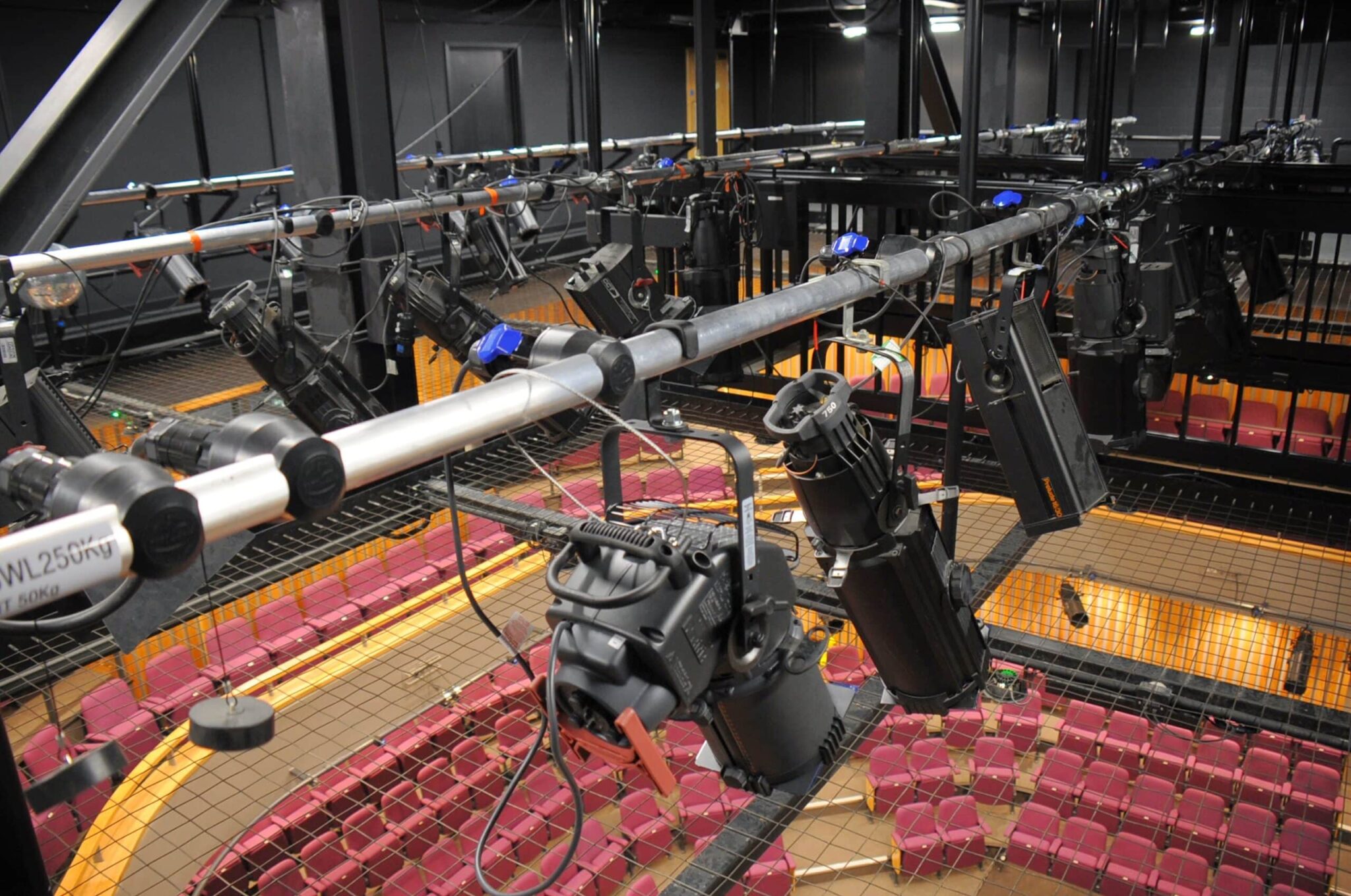 Stage lighting equipment and spotlights hang from metal bars above a theater with rows of red seats. A safety net covers the seating area below the lighting rig.