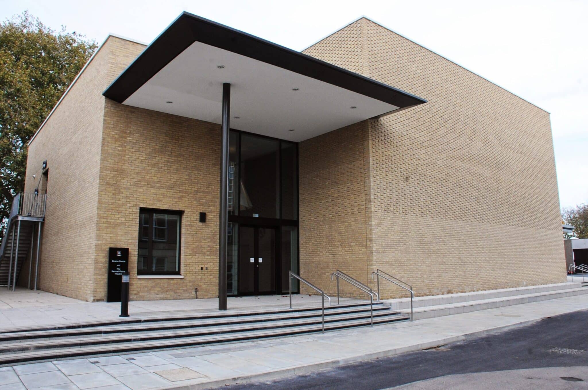 Modern brick building with large glass entrance, an overhanging flat roof, and metal railings along the wide steps leading to the doors. There is a black sign near the entrance and trees in the background.
