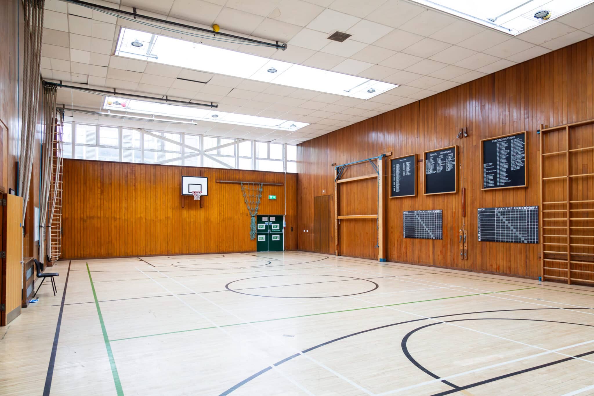 Empty indoor gymnasium with wooden walls, a basketball hoop, wall bars, climbing ropes, and skylights. The floor is marked with various sports lines. Plaques and equipment are mounted on the walls.