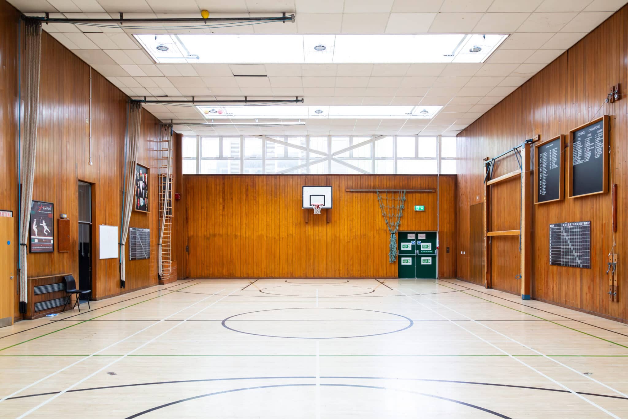 An empty indoor gymnasium with a basketball hoop, wooden paneled walls, sports markings on the light floor, wall-mounted scoreboards, benches, and a wall ladder. Large windows let in natural light from above.