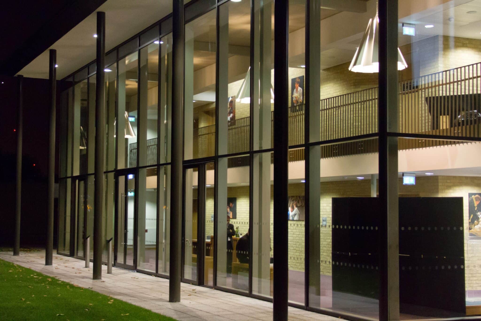 A modern building with large glass windows, interior lights illuminating a spacious lobby area with tables, chairs, and hanging lamps, viewed from outside at night.