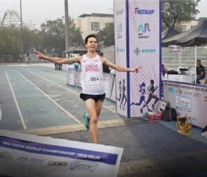 A male runner wearing a Great Britain vest and bib number 5014 crosses the finishing line with arms outstretched, smiling in victory, at the IAU 50km World Championship in New Delhi.