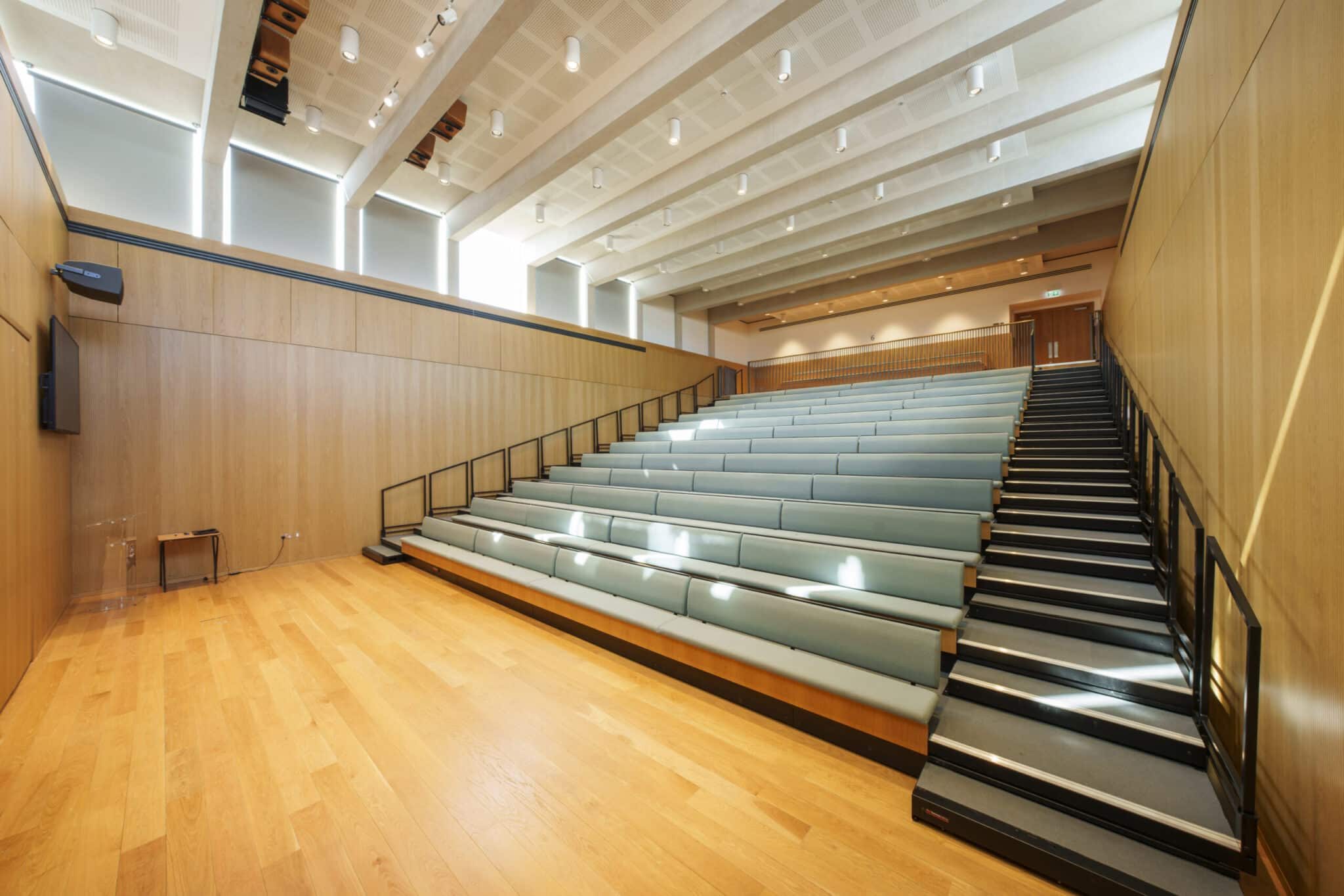 A modern lecture hall with wooden floors, light wood-paneled walls, tiered seating with green cushions, and black railings. Large windows near the ceiling let in natural light. The room is empty.