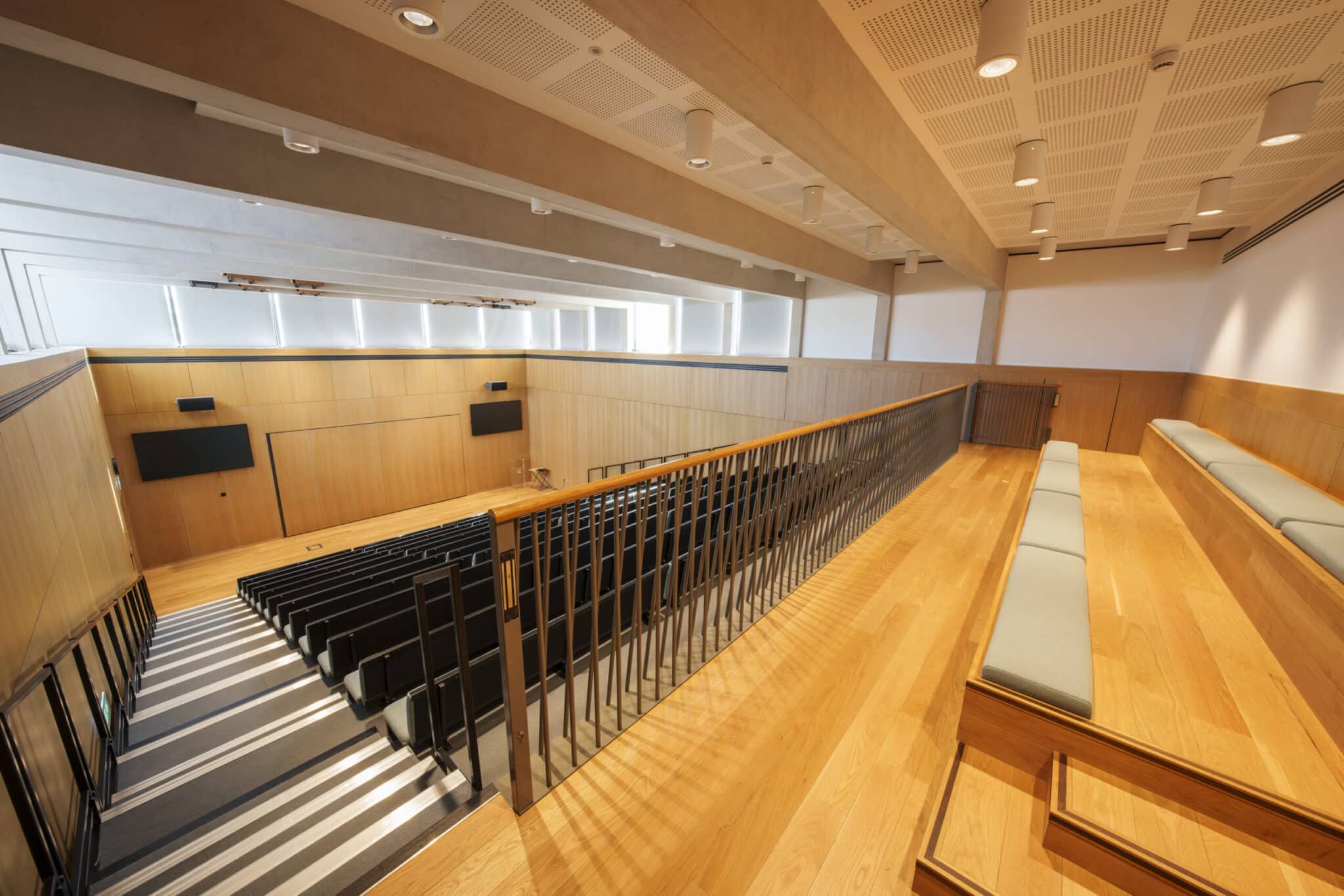 A modern lecture hall with wooden floors, tiered seating with black chairs on the left, and cushioned bench seating along the right side. The room has ample natural light and minimalistic design.