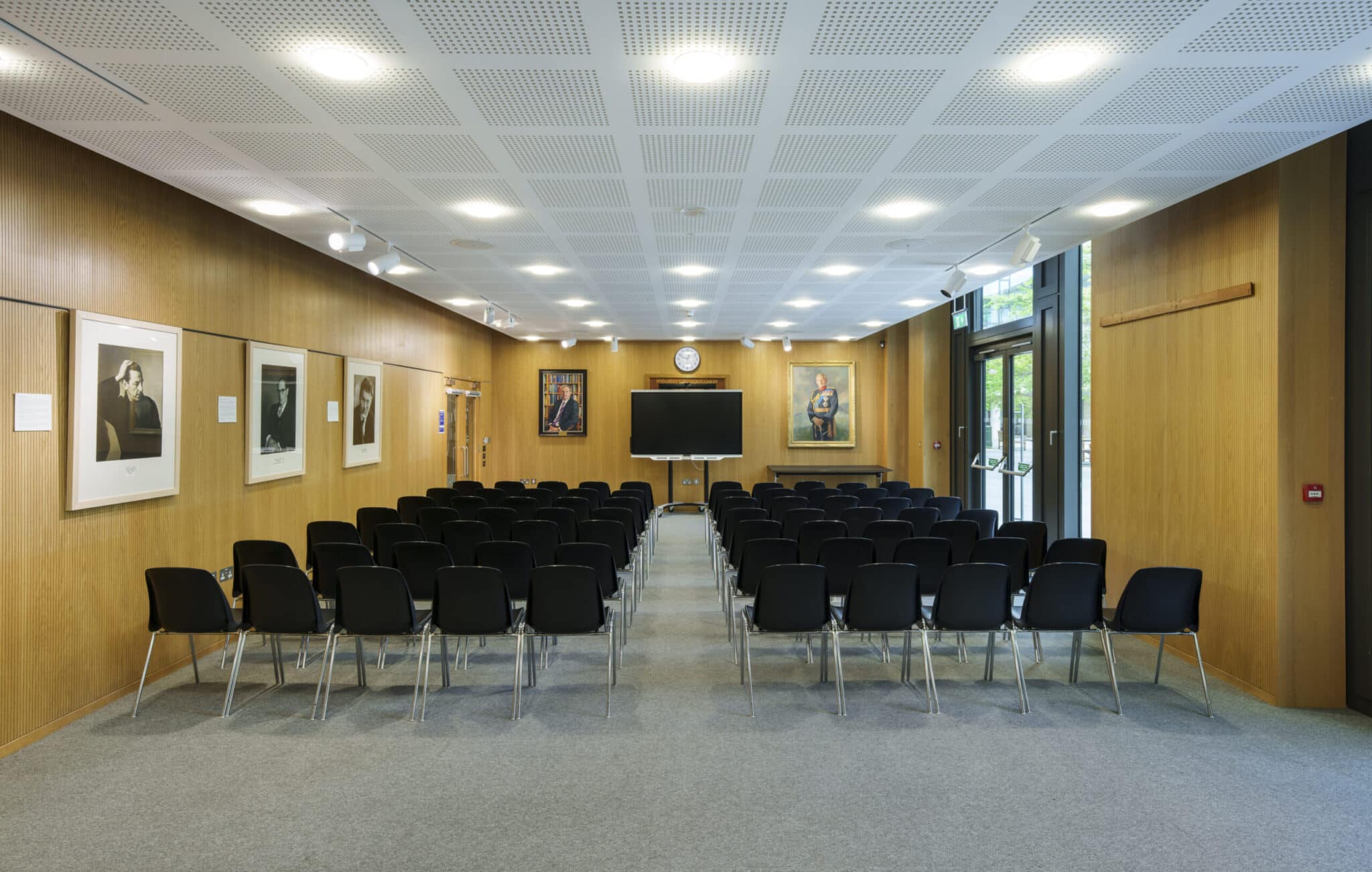 A conference room with rows of black chairs facing a large TV screen and clock on a wood-paneled wall. Framed portraits hang on the walls, and large windows let in natural light on the right side.