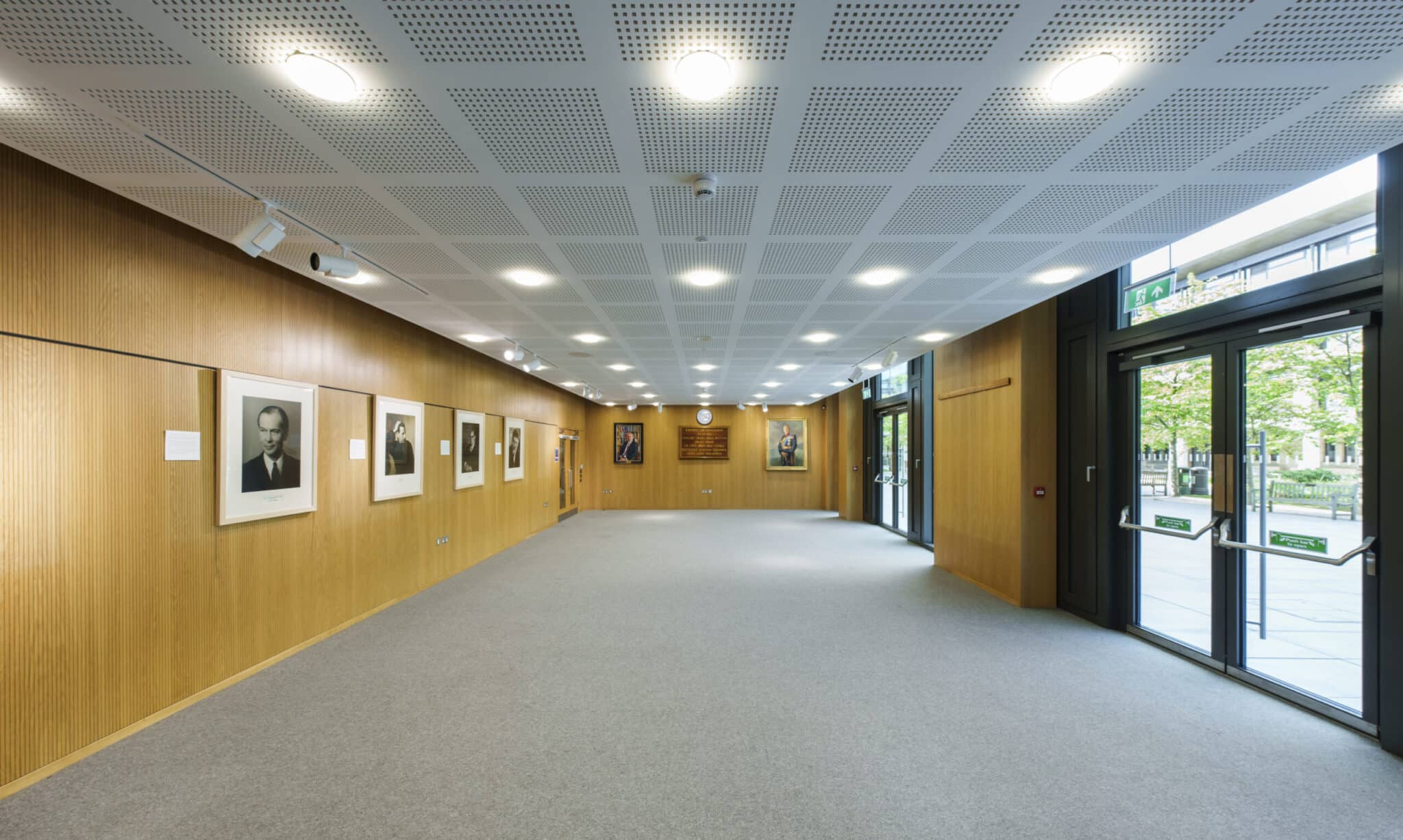 A spacious, well-lit hallway with wooden walls adorned with framed portraits and photographs. Large windows and glass doors on the right let in natural light, and the floor is carpeted in gray.