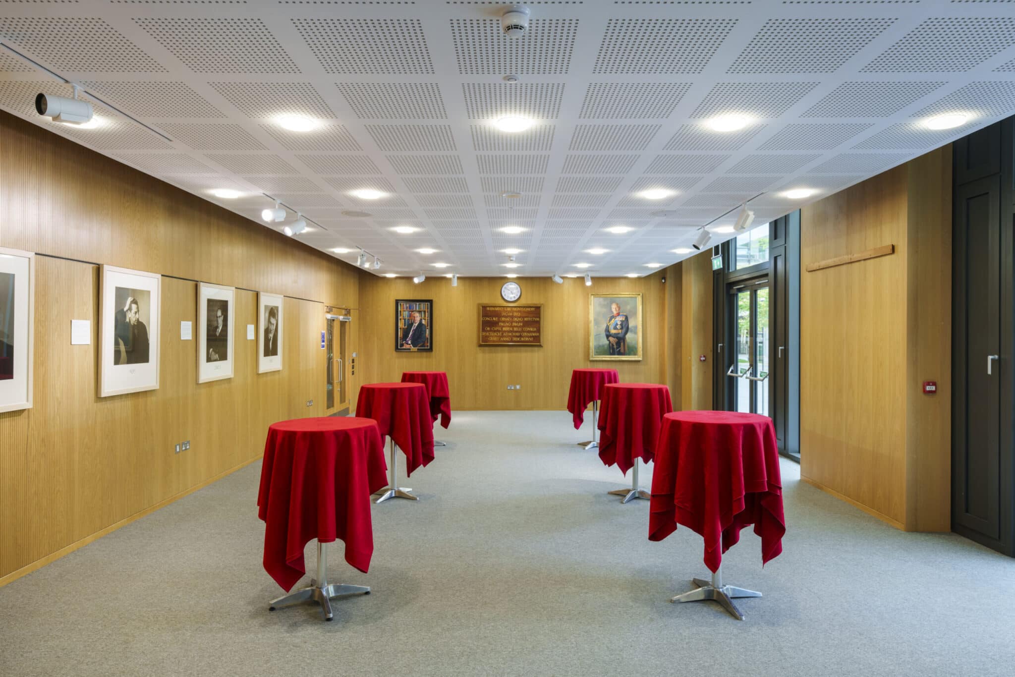 A bright, modern room with wood-paneled walls, portraits hanging, and several tall cocktail tables covered in red tablecloths spaced evenly across a gray carpeted floor. Large windows line one side.