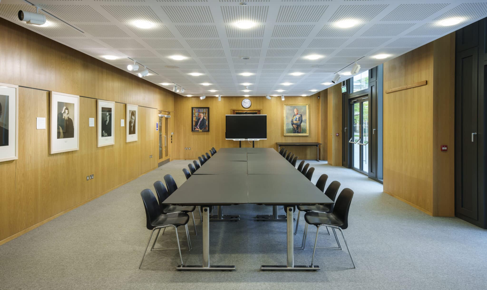 A modern conference room with a long black table surrounded by black chairs, wood-paneled walls, framed portraits, a large monitor, and windows letting in natural light.