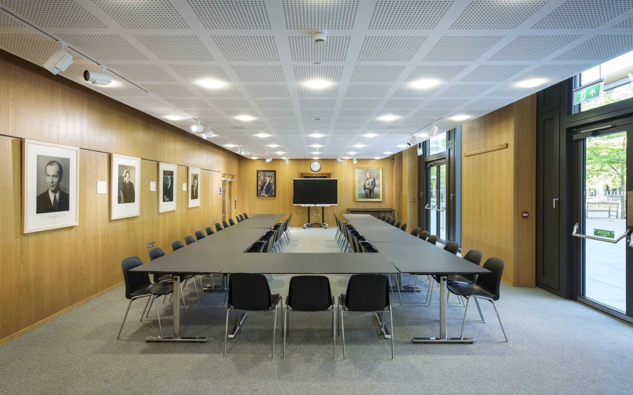 A modern conference room with a U-shaped table arrangement, black chairs, wood-paneled walls, portraits on the walls, a large screen at the front, and large windows letting in natural light.