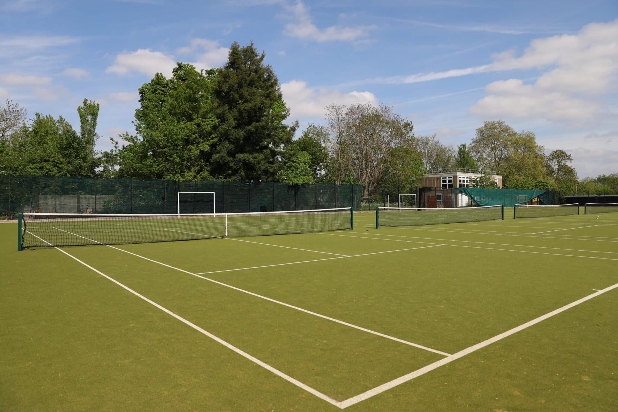 An outdoor tennis court with a green surface and white lines, surrounded by trees and a fence, with a building visible in the background under a partly cloudy sky.