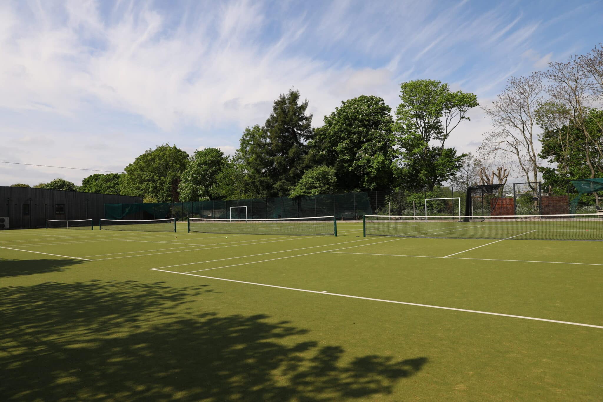 A green outdoor tennis court with white lines sits empty under a partly cloudy sky, surrounded by trees and a netted fence. Shadows from nearby trees fall across the court.
