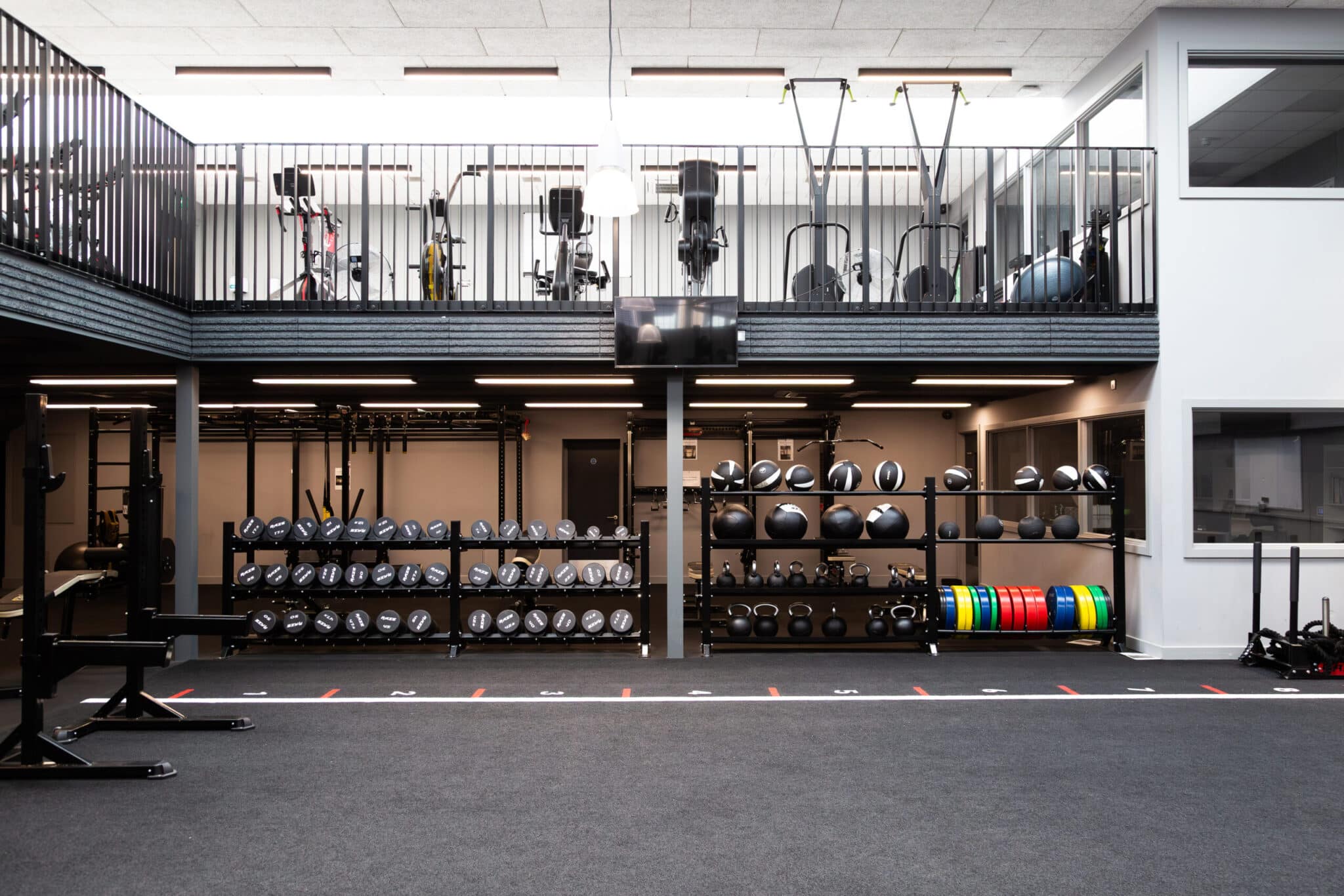Modern gym with racks of medicine balls, weight plates, benches, and exercise equipment. There is a mezzanine level with cardio machines and railings, and the space is well-lit with natural light from windows.