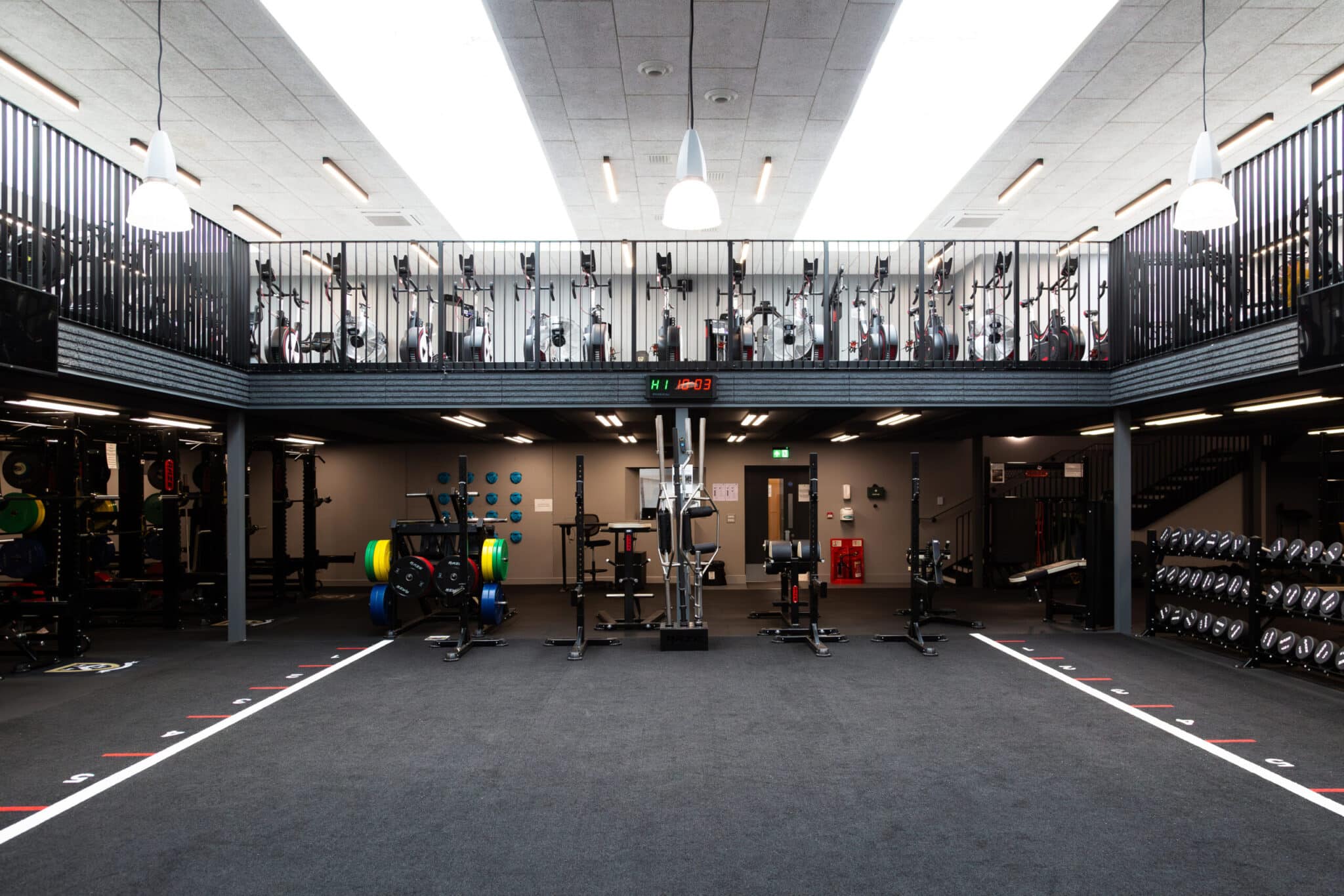 Modern gym interior with workout equipment, weights, and machines neatly organized. There is an upper mezzanine level with exercise bikes and the area is well-lit with overhead lights. The floor is spacious and marked for workouts.