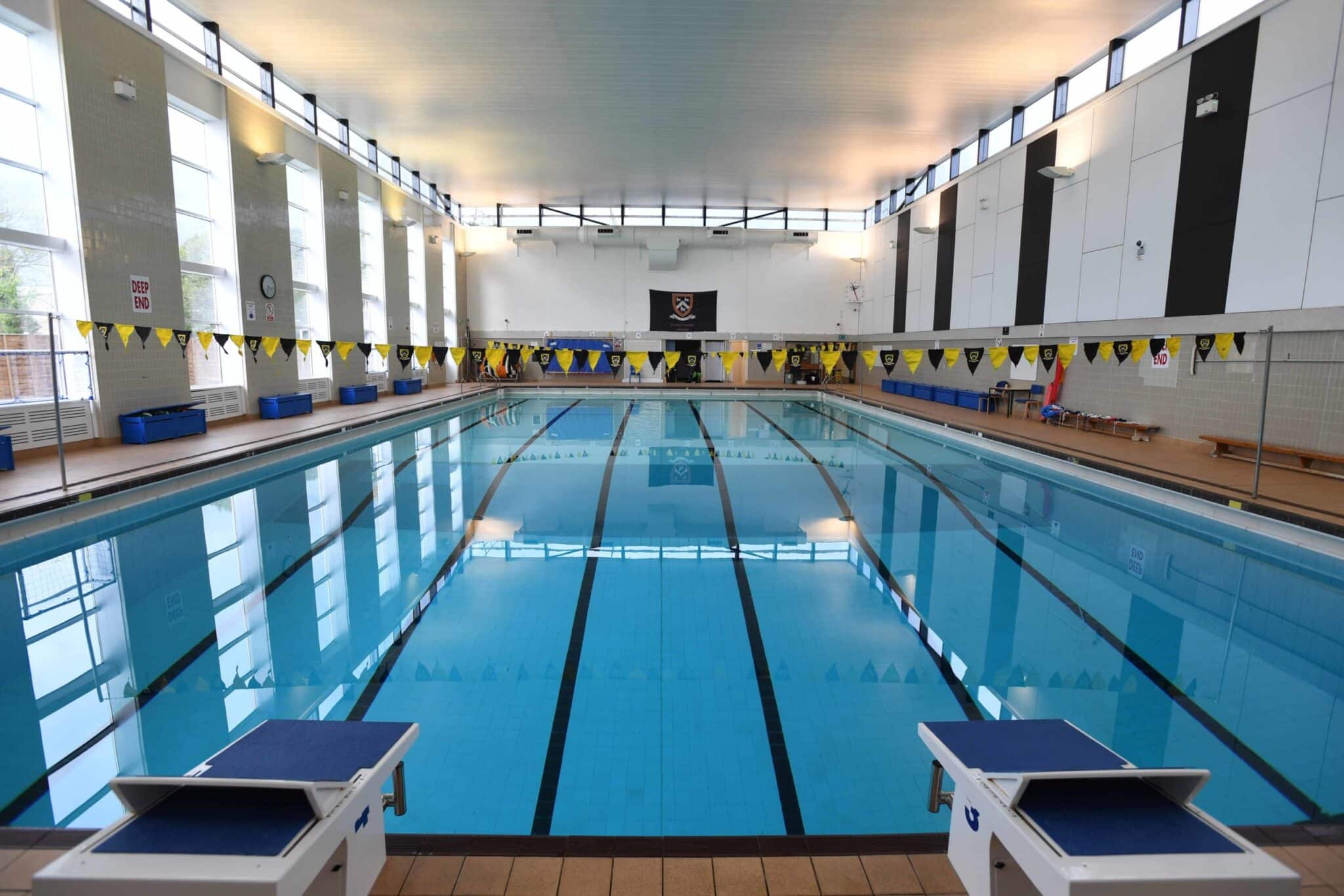 Indoor swimming pool with clear blue water, lane markings, starting blocks, and yellow triangular flags overhead. Large windows line the sides, allowing natural light into the spacious, empty pool area.