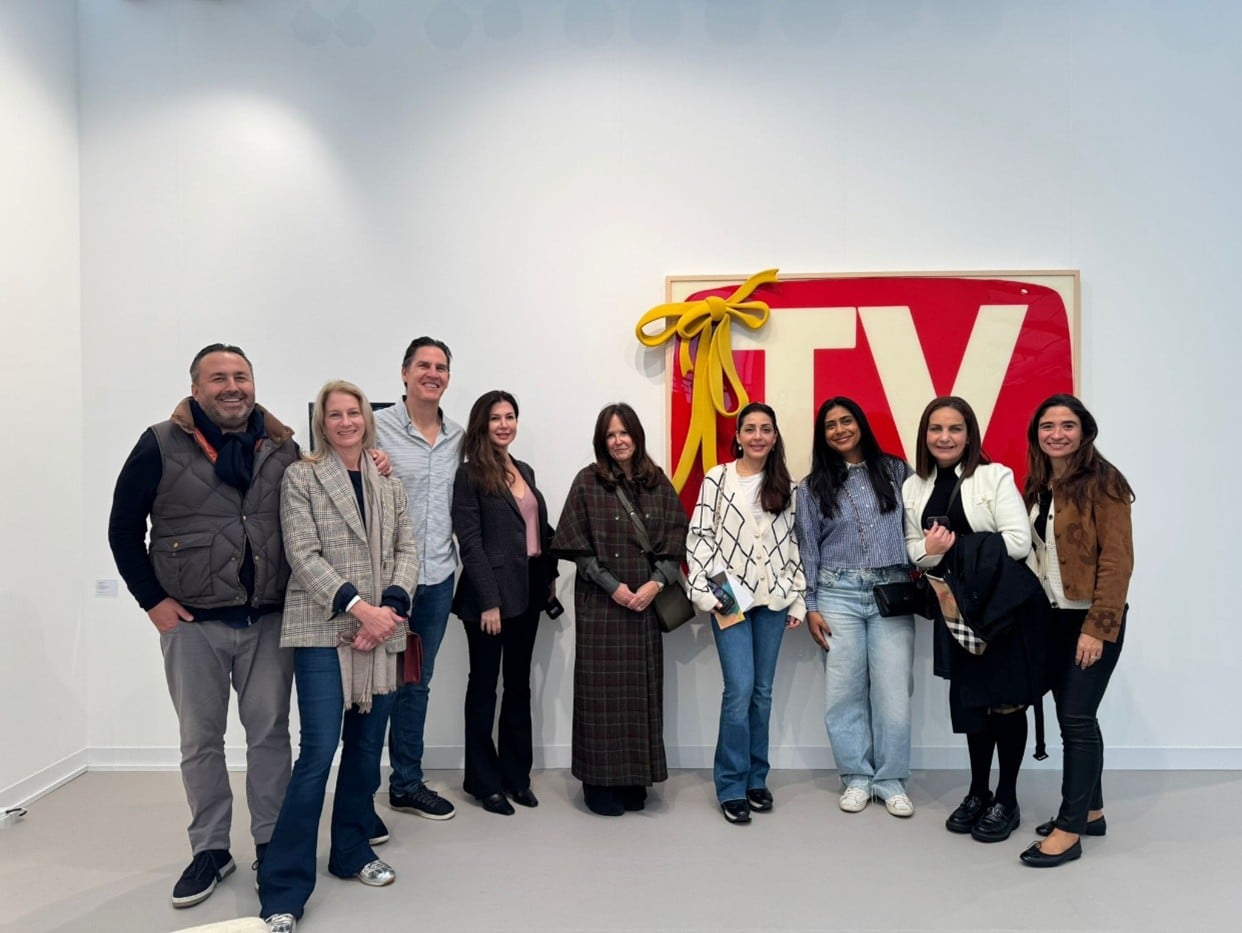 A group of nine people stand smiling in front of a wall art piece resembling a wrapped gift with the letters TV in red and a large yellow bow. The group is casually dressed and posing indoors.