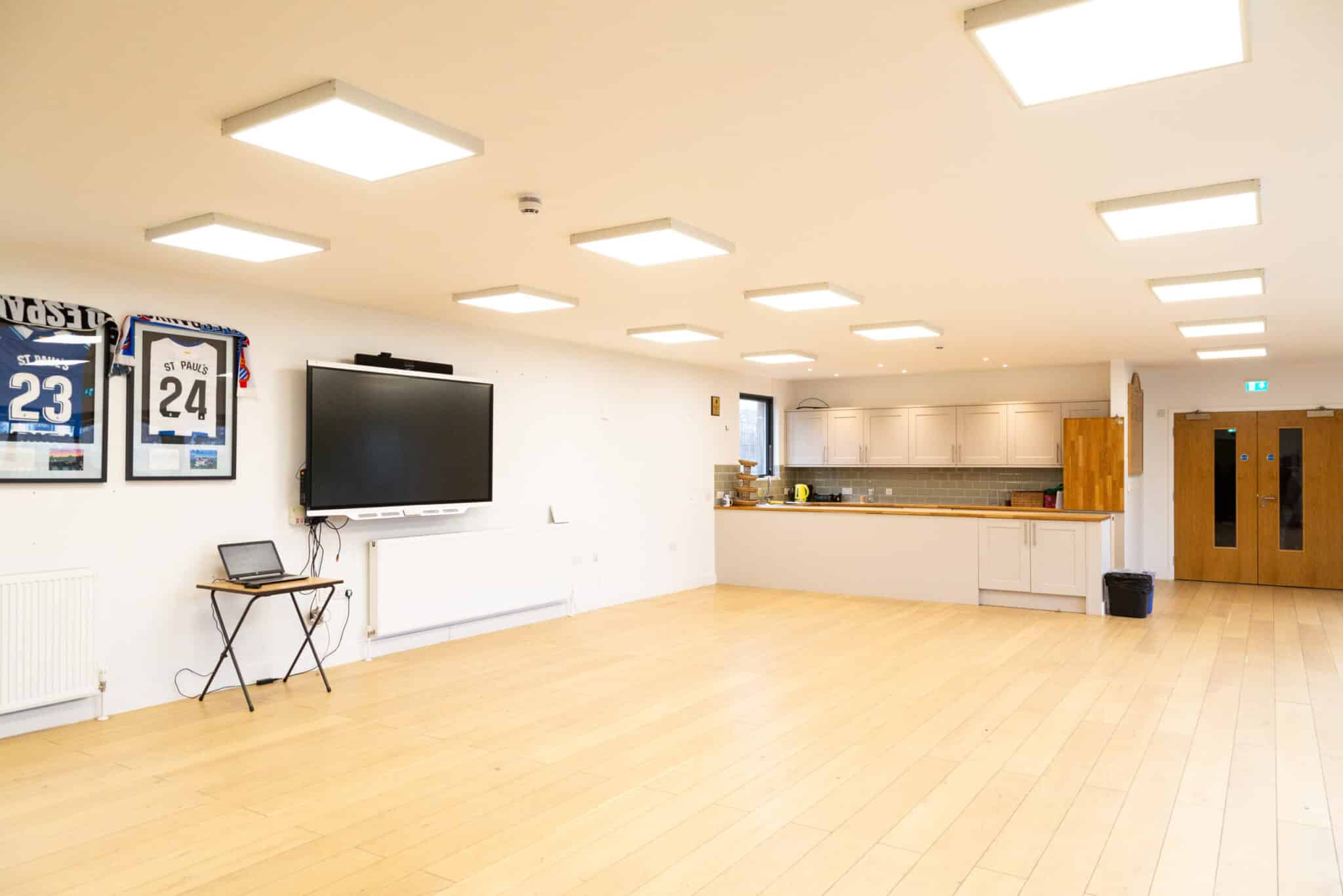 A spacious, well-lit room with wooden floors, a small kitchen area in the back, a wall-mounted TV, two framed sports jerseys, a folding table with a laptop, and double wooden doors on the right.