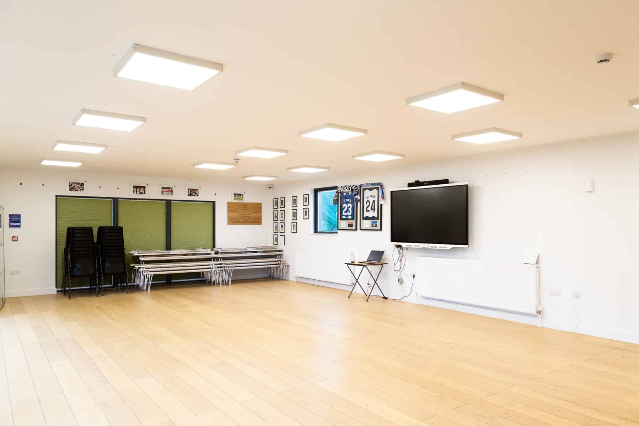 A spacious, well-lit room with wooden flooring, stacked chairs and tables along one wall, framed jerseys and photos on another, a large TV screen, and a small table with a laptop.