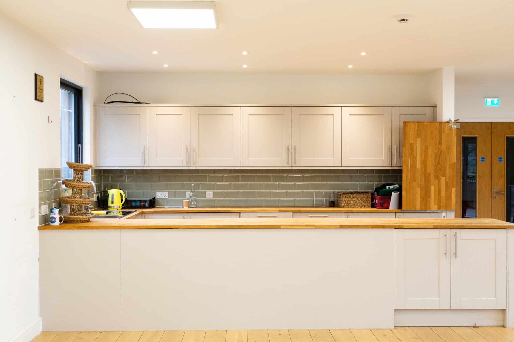 Modern kitchen with white cabinets, light wood countertops, green subway tile backsplash, a wooden floor, a stacked basket stand, a yellow kettle, and various items neatly placed on the counter and shelves.