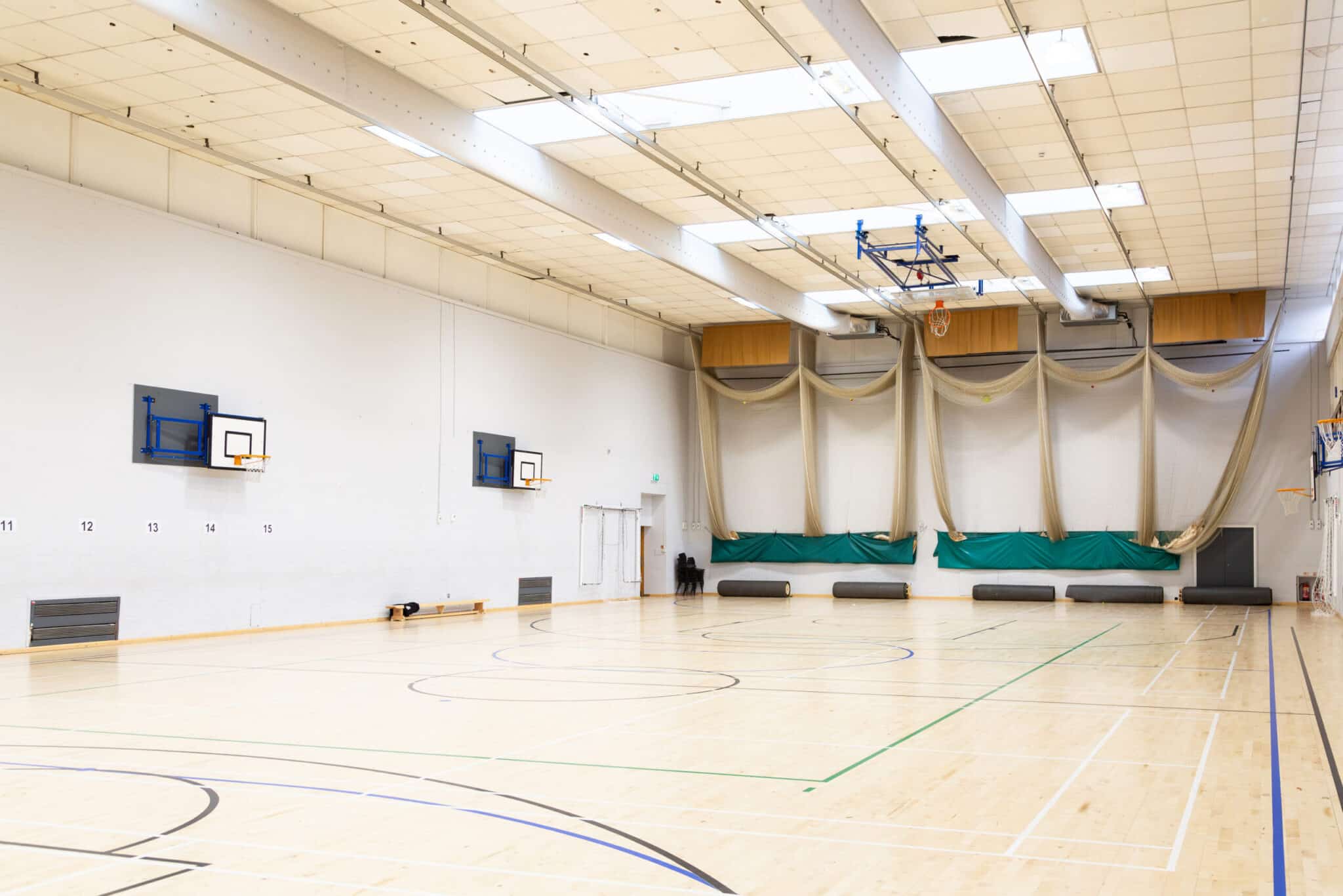 Brightly lit, empty indoor gymnasium with a light wood floor, basketball hoops on the walls, green and white curtains, and benches along the side. The ceiling has exposed beams and lighting fixtures.