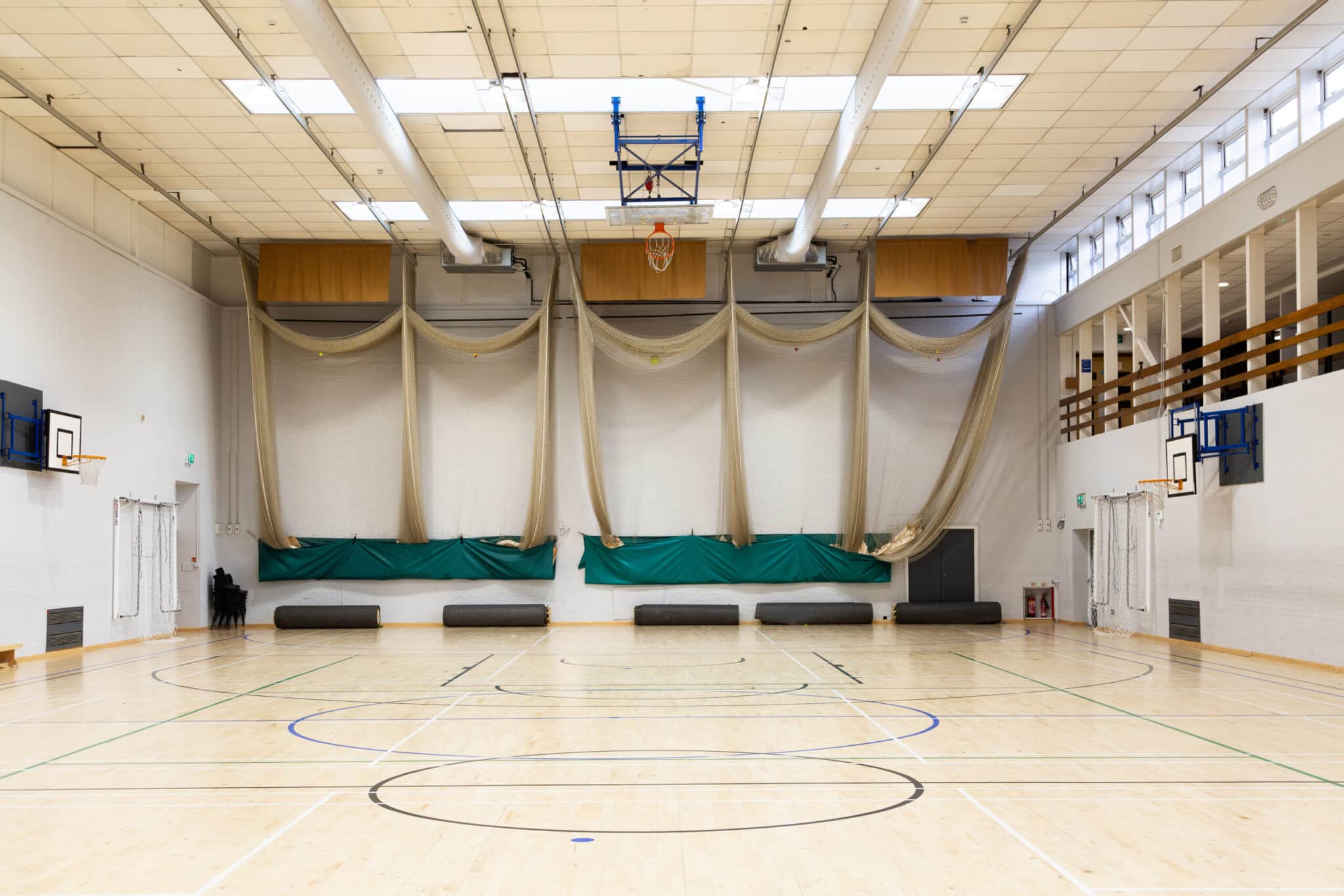 Empty indoor gymnasium with a wooden floor, basketball hoops, netting hanging on the far wall, large windows on the right, and benches along the walls. Overhead lights and exposed pipes are visible on the ceiling.