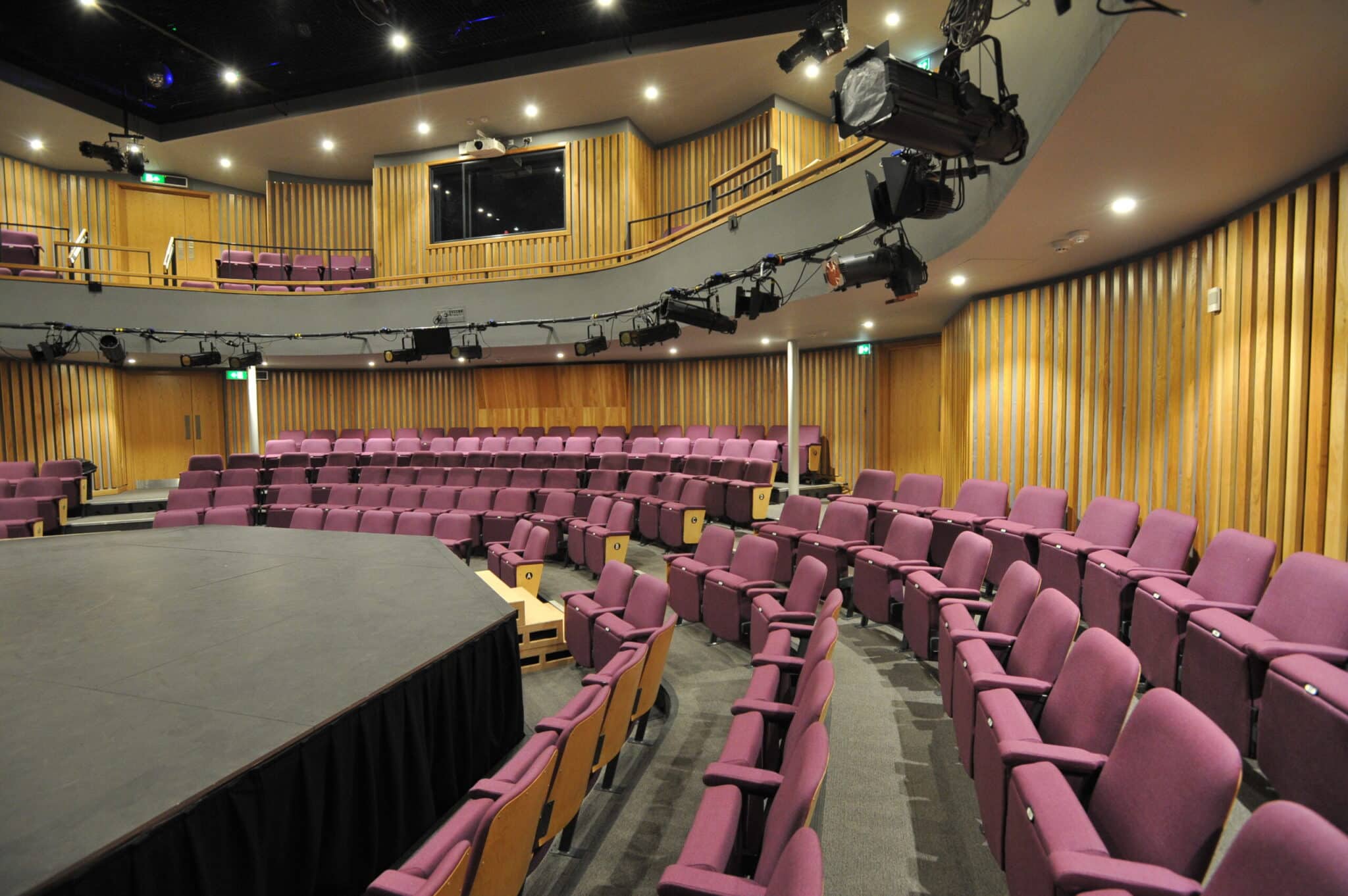 A modern theater with rows of purple seats arranged in a semi-circle around a central stage, wooden panel walls, overhead lighting, and a balcony with additional seating.