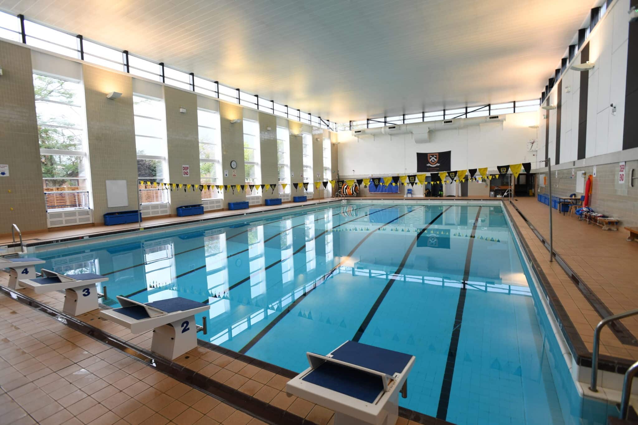 Indoor swimming pool with clear blue water, starting blocks, lane markings, and yellow-and-black flags overhead. Large windows allow natural light in, and the pool area is surrounded by tiled flooring and benches.