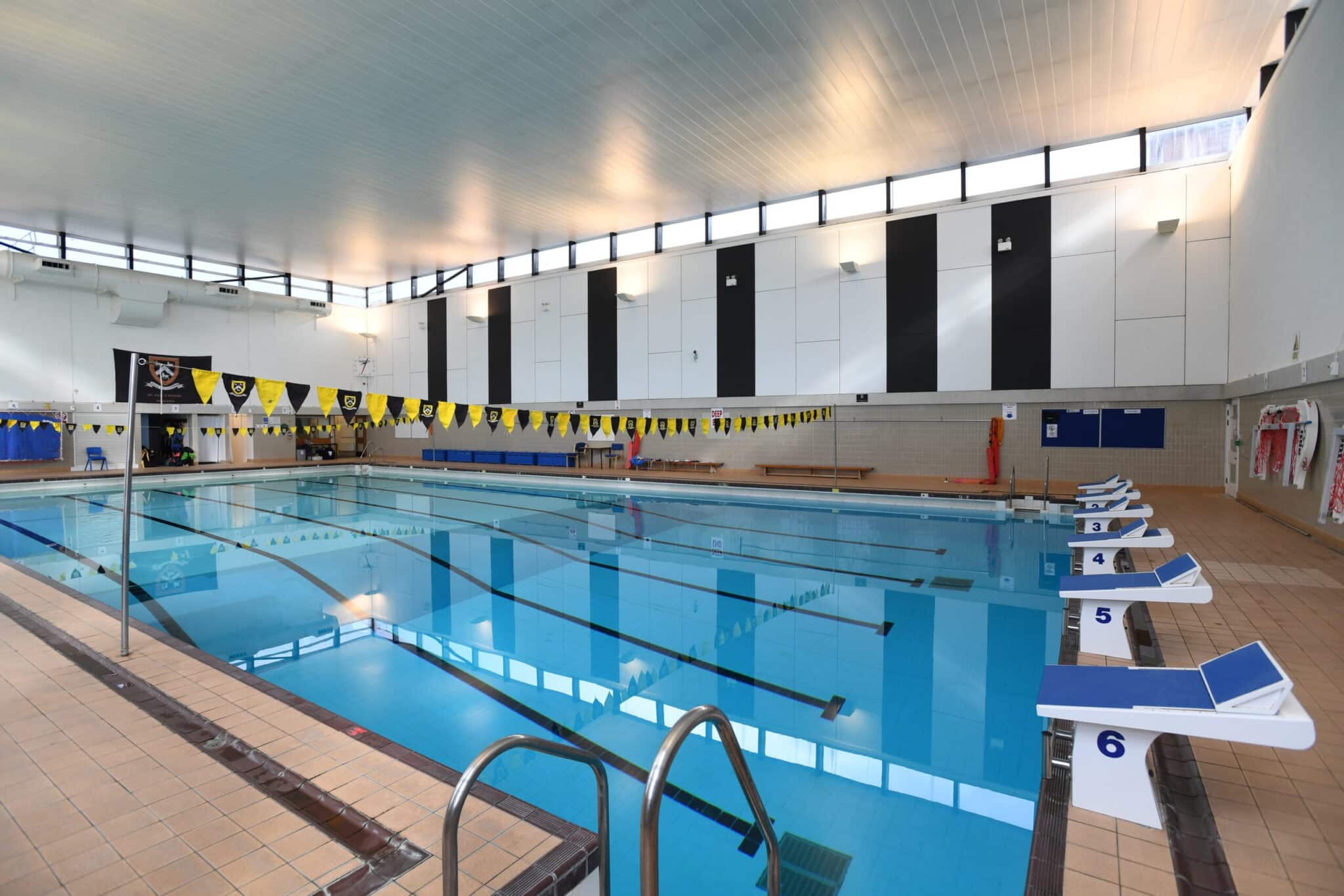 Indoor swimming pool with six swim lanes, starting blocks, lane dividers with yellow and black flags, tiled deck, benches along the wall, and large windows letting in natural light.
