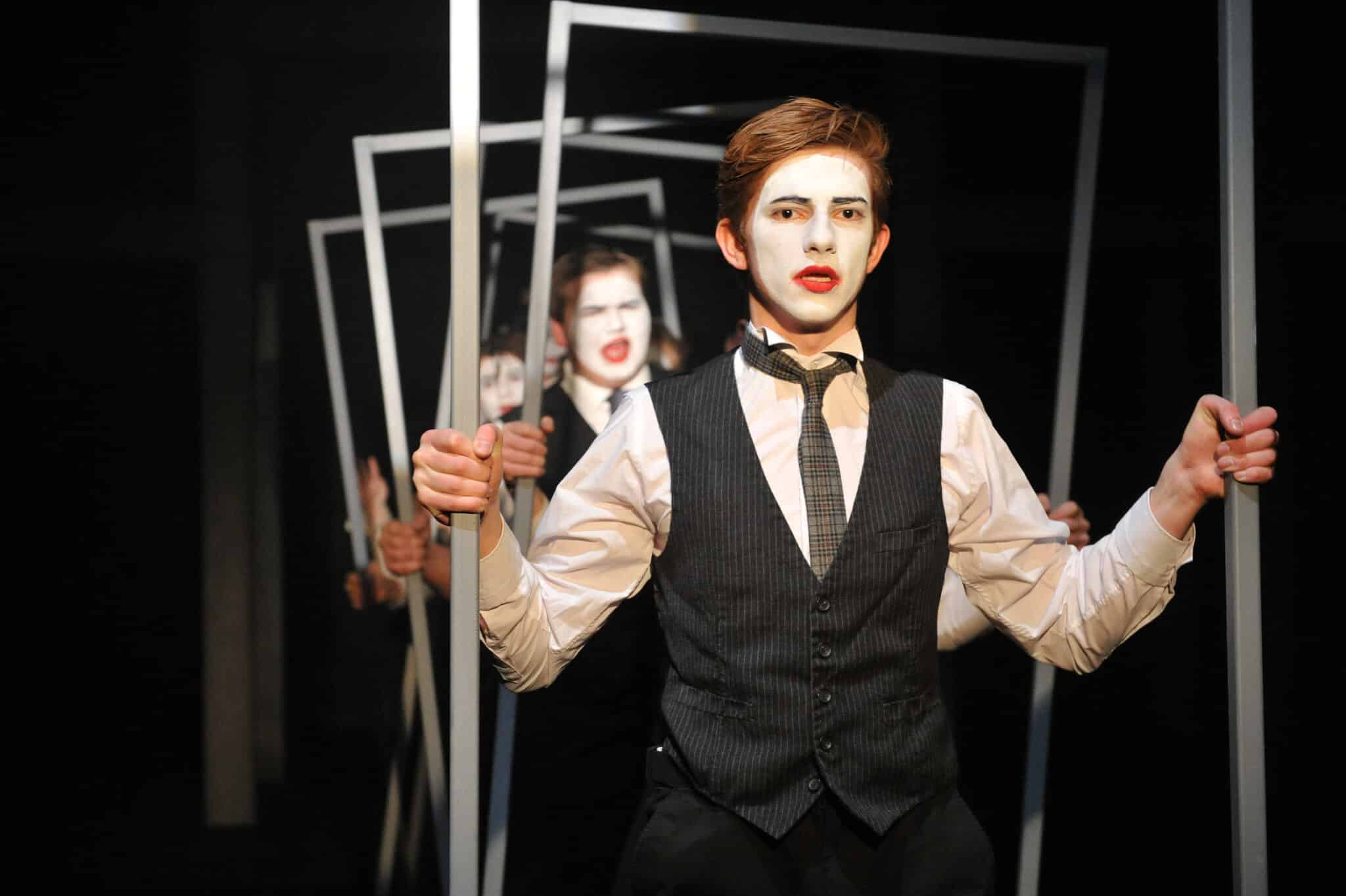A person wearing white face paint, red lipstick, a suit vest, and tie holds a rectangular frame. Others dressed similarly stand behind, each framed, creating a layered, theatrical effect.