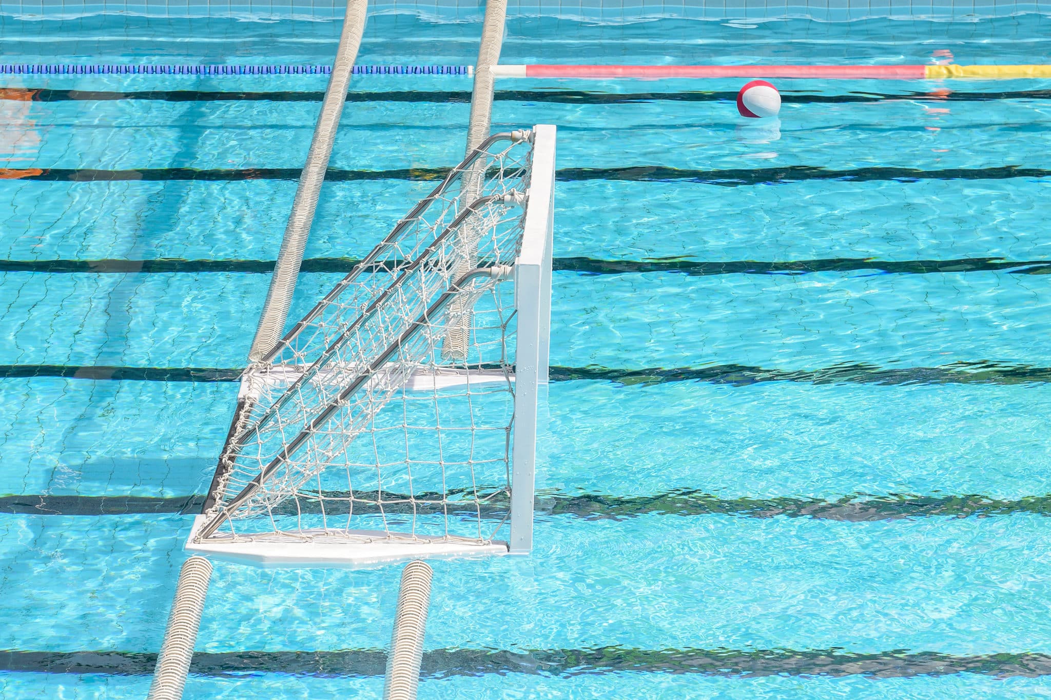 A floating water polo goal is positioned in a clear blue swimming pool. A red and white water polo ball floats on the water near colorful lane markers. Black lane lines are visible on the pool floor.