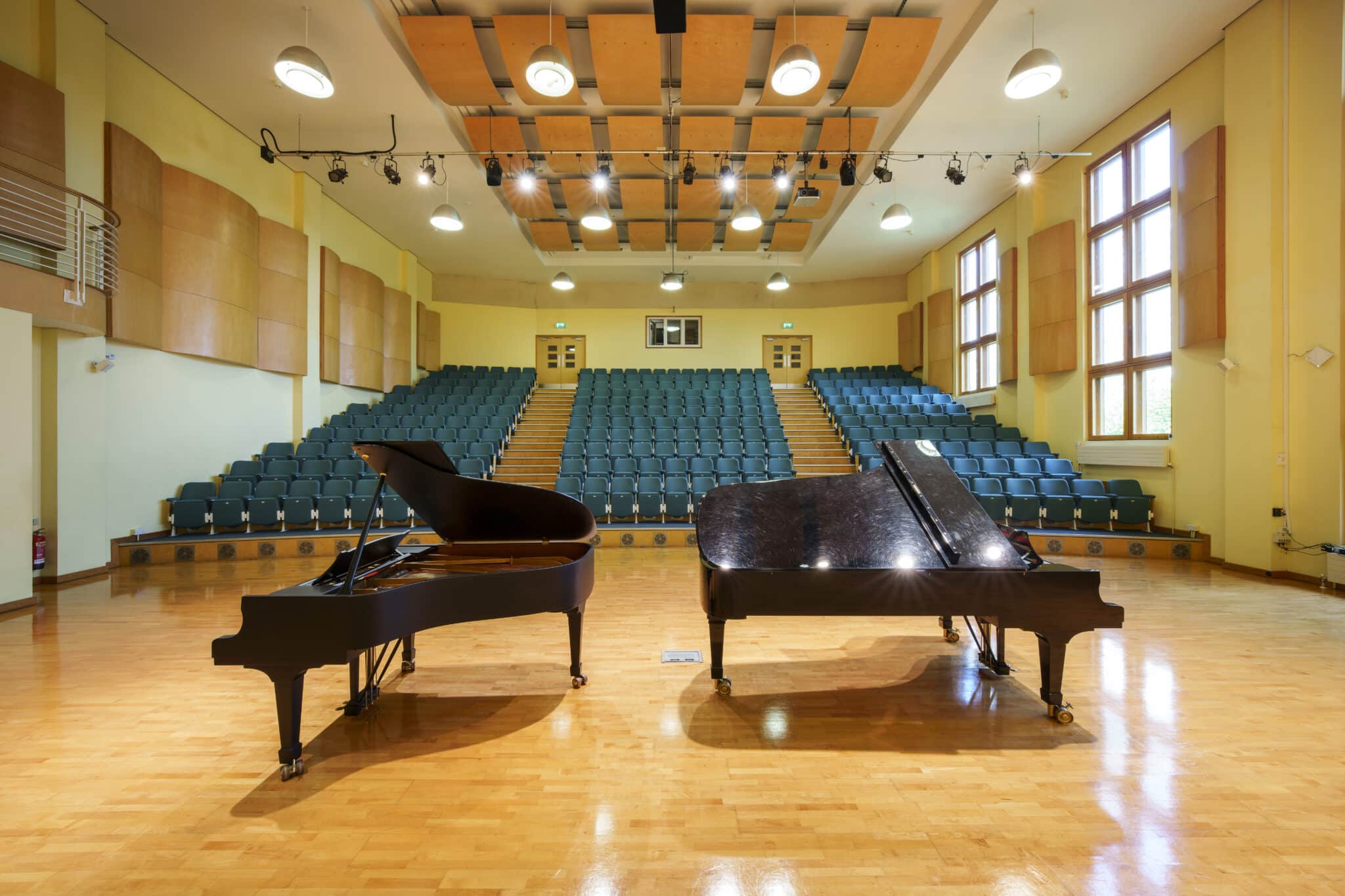 Two grand pianos are placed side by side on a polished wooden floor in a spacious, empty concert hall with rows of green seats and large windows letting in natural light.