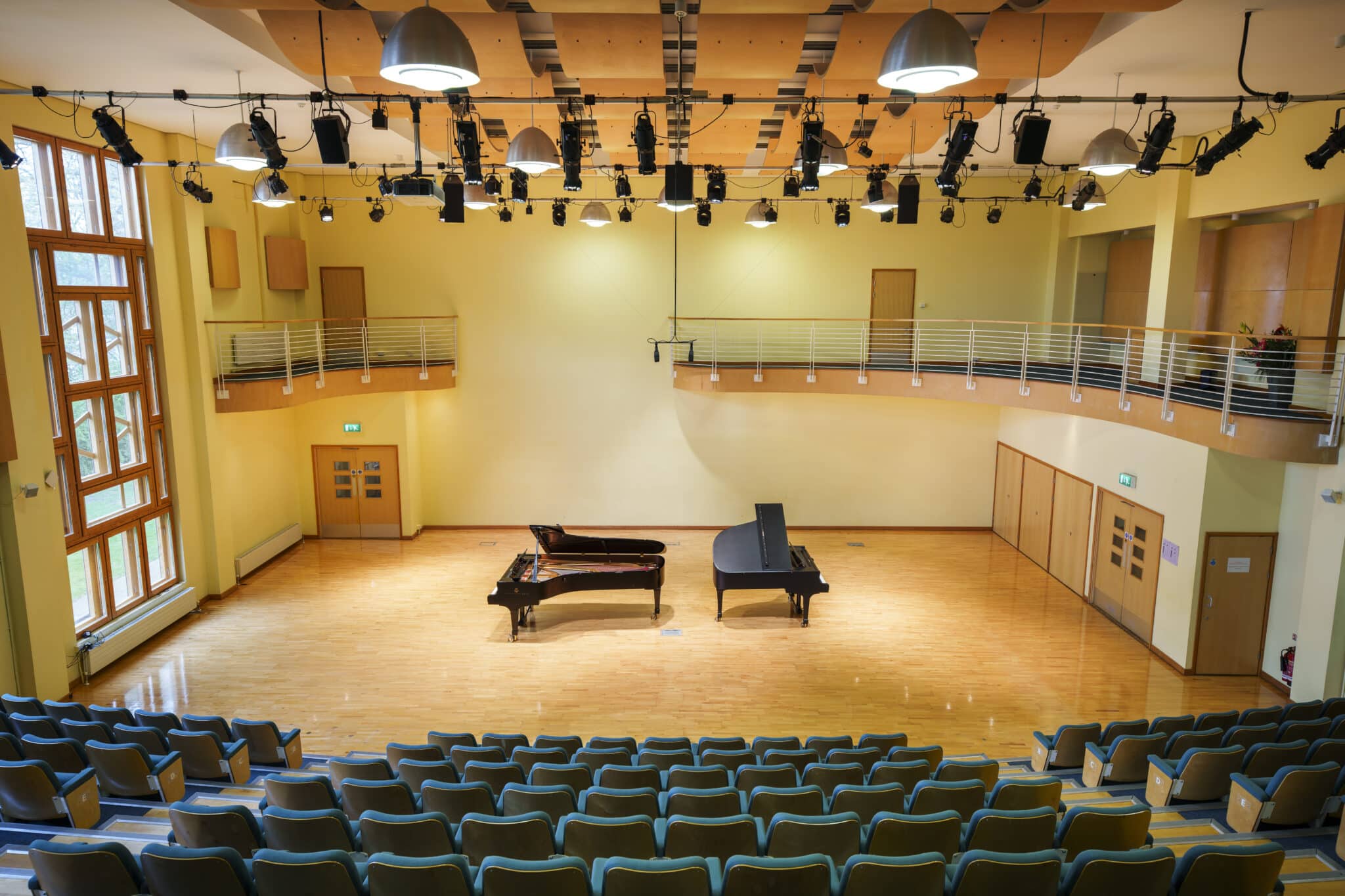 A concert hall with rows of empty seats facing a stage where two grand pianos are positioned. The hall has wooden floors, yellow walls, large windows, and a balcony with railings above the stage area.