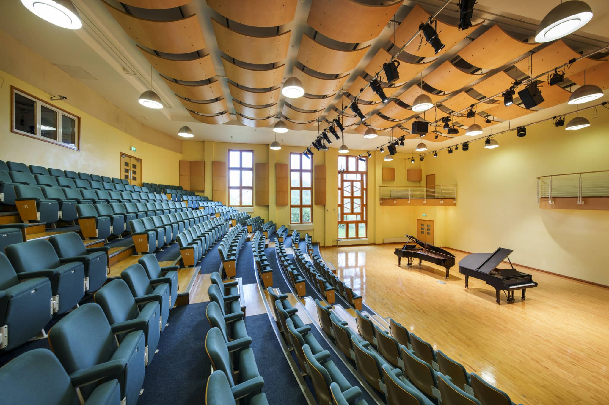 A modern auditorium with tiered seating, wooden floors, large windows letting in natural light, and two grand pianos on stage. Curved acoustic panels hang from the ceiling, and stage lighting is arranged above.