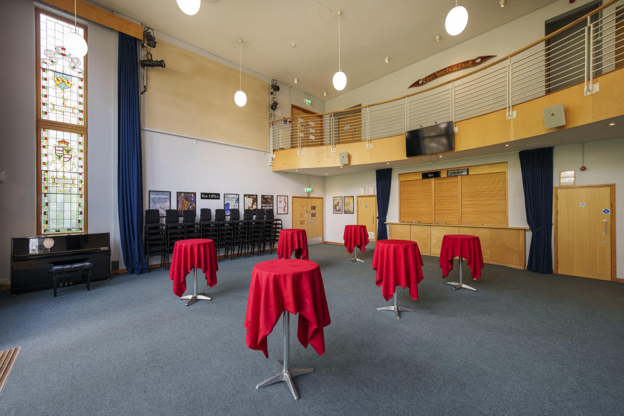 Spacious room with high ceilings, stained glass windows, and a mezzanine. Six small tables with red tablecloths are evenly spaced, and stacked chairs line one wall. The decor is modern with theatre posters displayed.