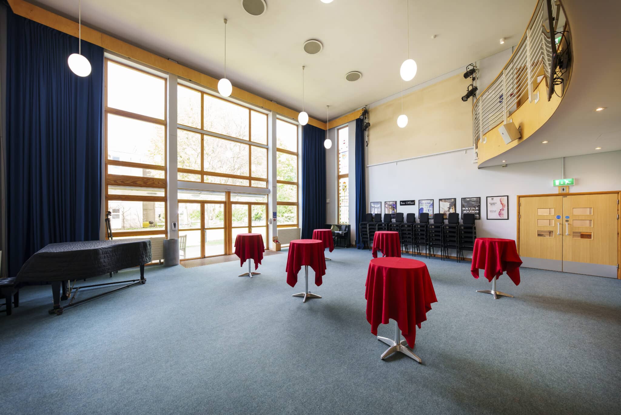 A spacious, modern event room with tall windows, round tables covered in red cloth, stacked chairs in the corner, and hanging globe lights. Blue curtains frame the windows and a balcony overlooks the space.