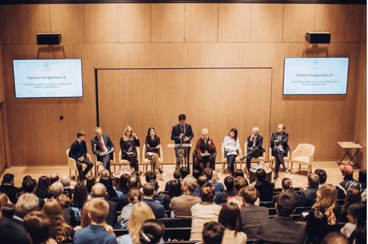 A panel of eight people sits onstage in a lecture hall with an audience facing them; one person stands at a podium speaking, and two screens display the text Pauline Perspectives X.