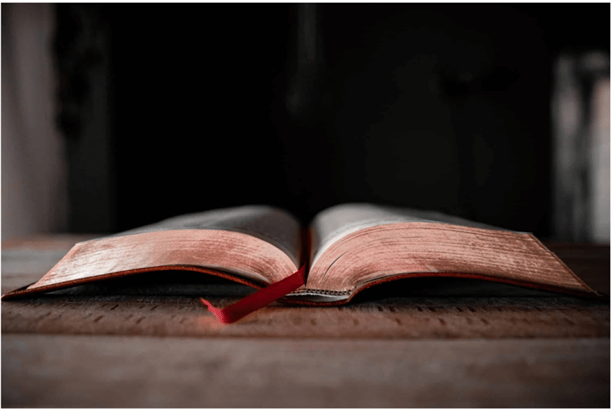An open book with a red ribbon bookmark lying on a wooden surface, with a dark and blurred background.